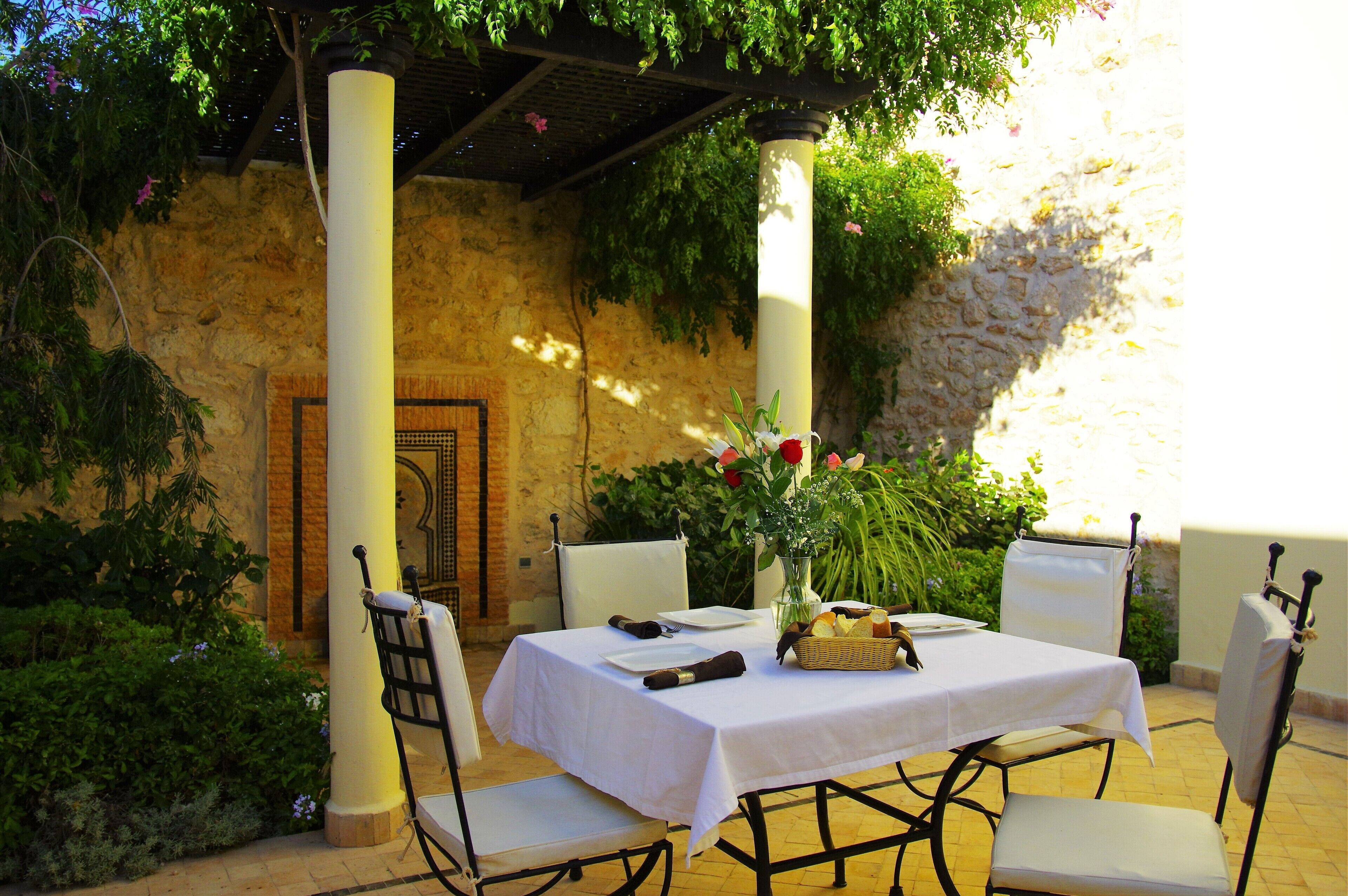 Un espace de restauration en plein air avec une table dressée pour deux personnes, entourée de chaises blanches, sous une pergola couverte de plantes grimpantes.