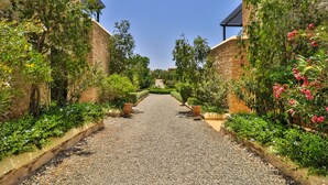 Un sentier bordé d’une végétation luxuriante et de murs de pierre, qui mène à un bâtiment situé au loin.