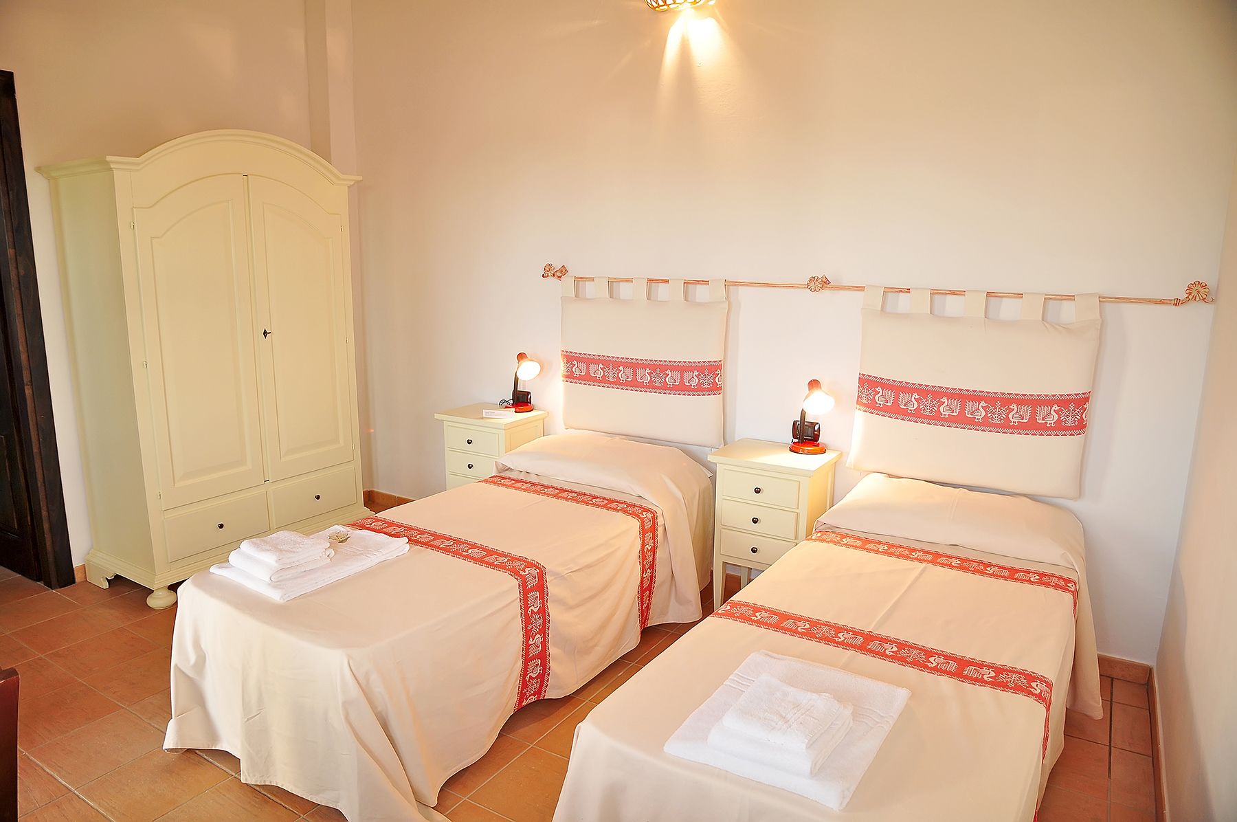 Habitación de hotel con dos camas, cada una con una colcha estampada en rojo y blanco, una mesita de noche con lámpara y un armario.