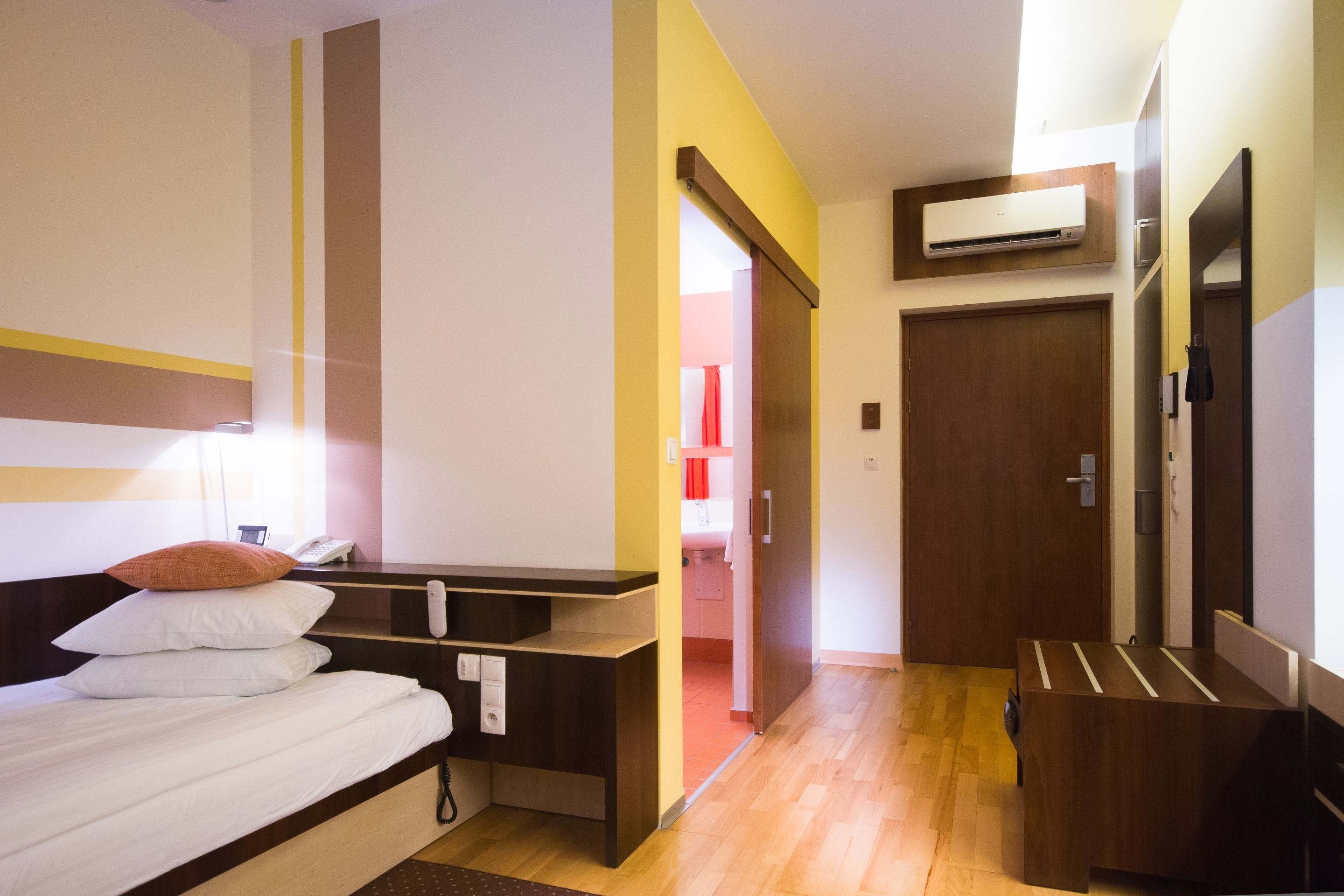 Camera d'albergo con un letto, comodino e una scrivania. Si apre una porta che dà su un bagno e un corridoio.