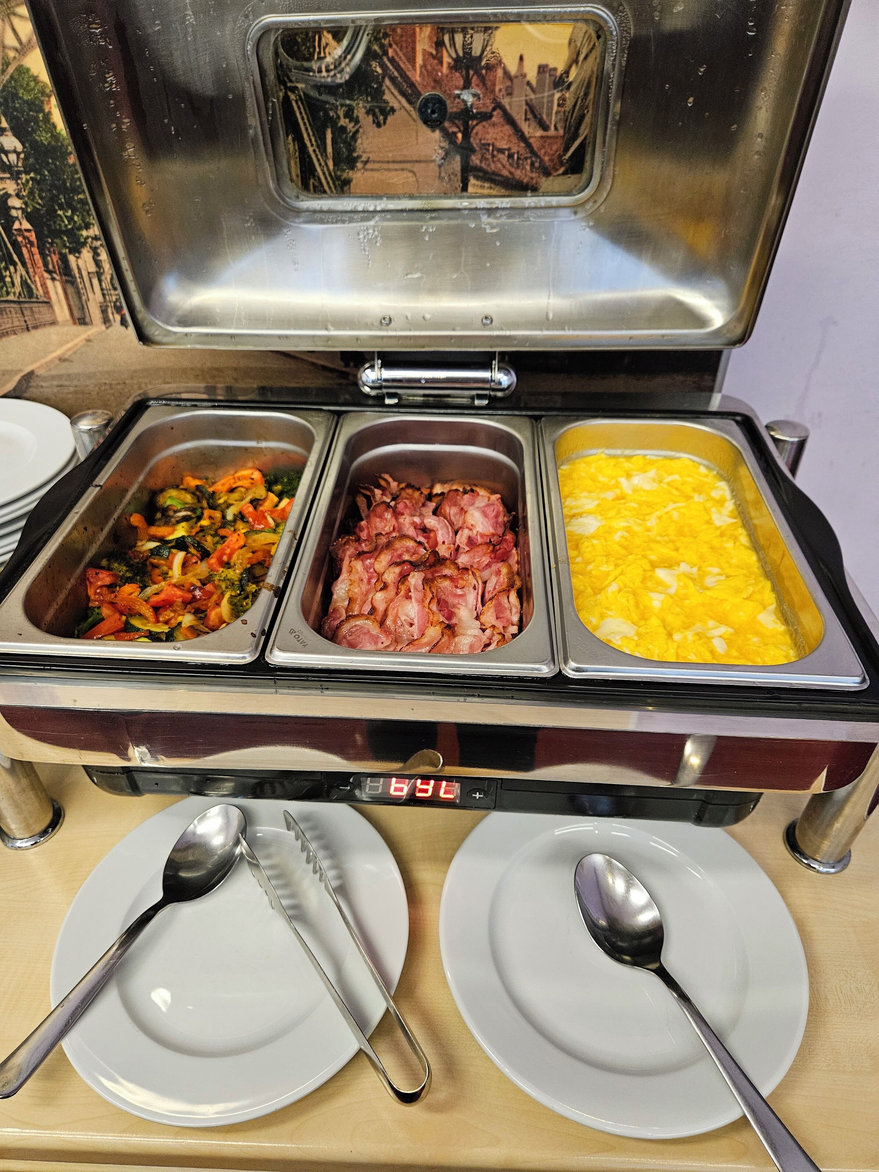 Ein Buffet mit drei Behältern: einer mit gemischtem Gemüse, einer mit Speck und einer mit Rührei. Auf einem Holztisch stehen zwei weiße Teller mit Löffeln.