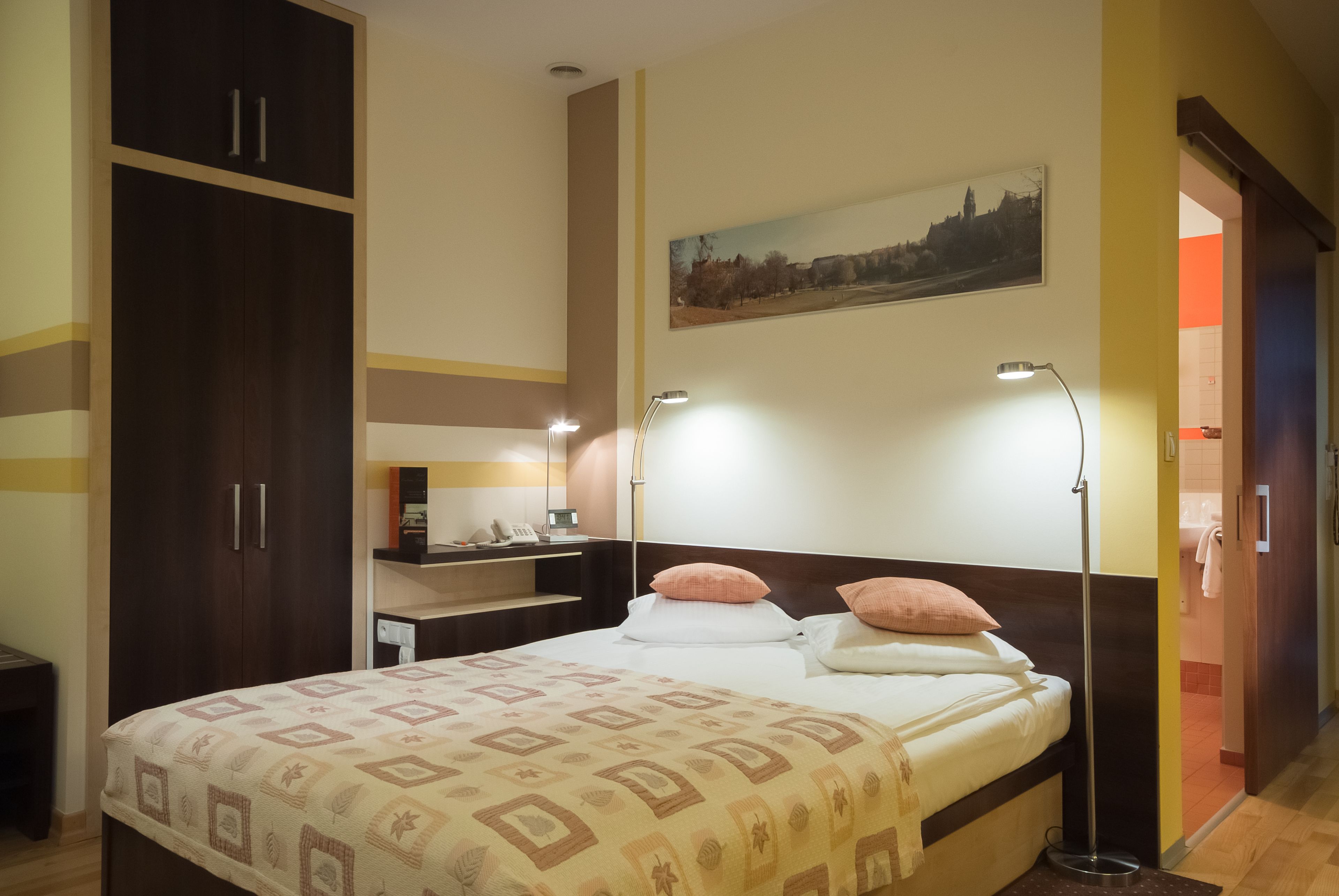 Habitación de hotel con cama, lámparas de noche, armario, escritorio y un cuadro en la pared.