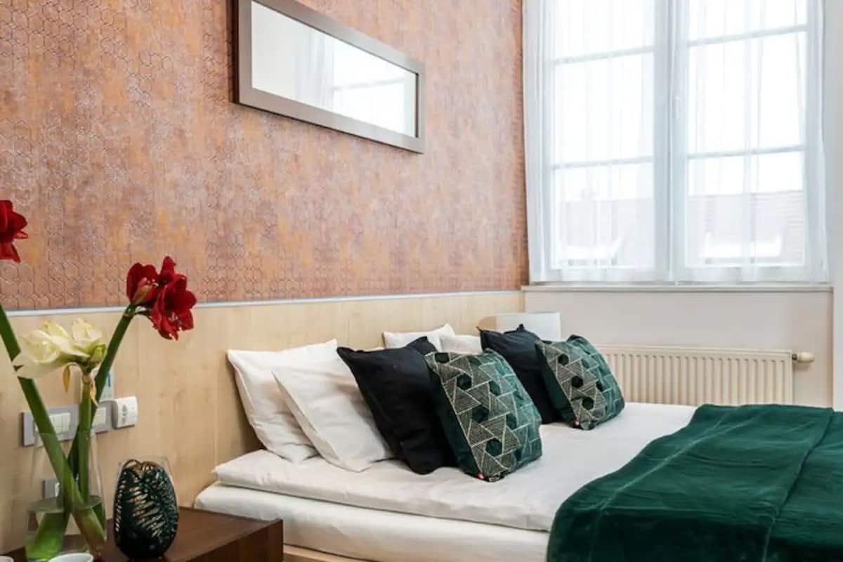 Quarto de hotel com uma cama, almofadas, mesa de cabeceira com um vaso de flores e uma janela com persianas.