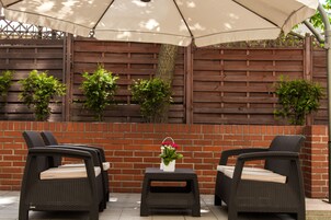 An outdoor seating area with wicker chairs and a table, under a large umbrella, against a brick wall with plants.