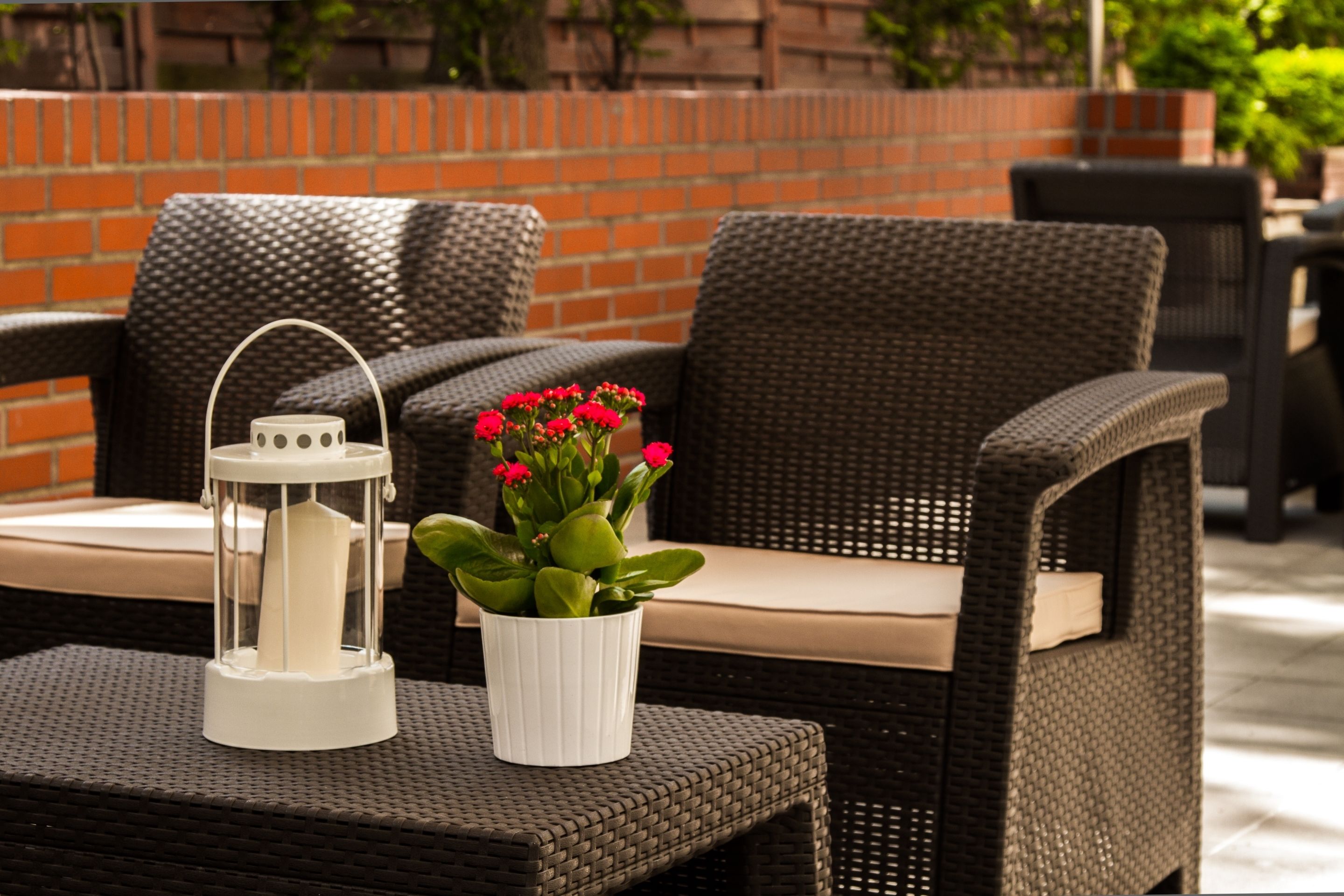 Korbmöbel mit Kissen, ein weißer Laternen und ein Blumentopf mit roten Blüten.