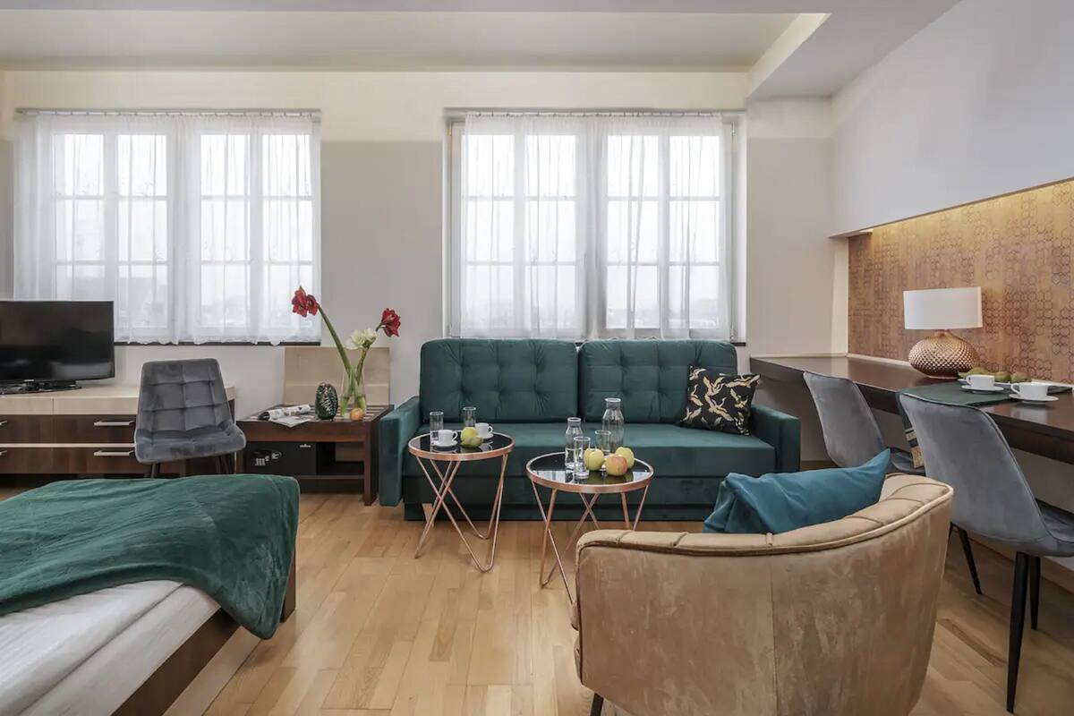 Quarto de hotel moderno com sofá verde, uma cama, uma escrivaninha com televisão, duas mesinhas redondas com bebidas e um abajur decorativo.