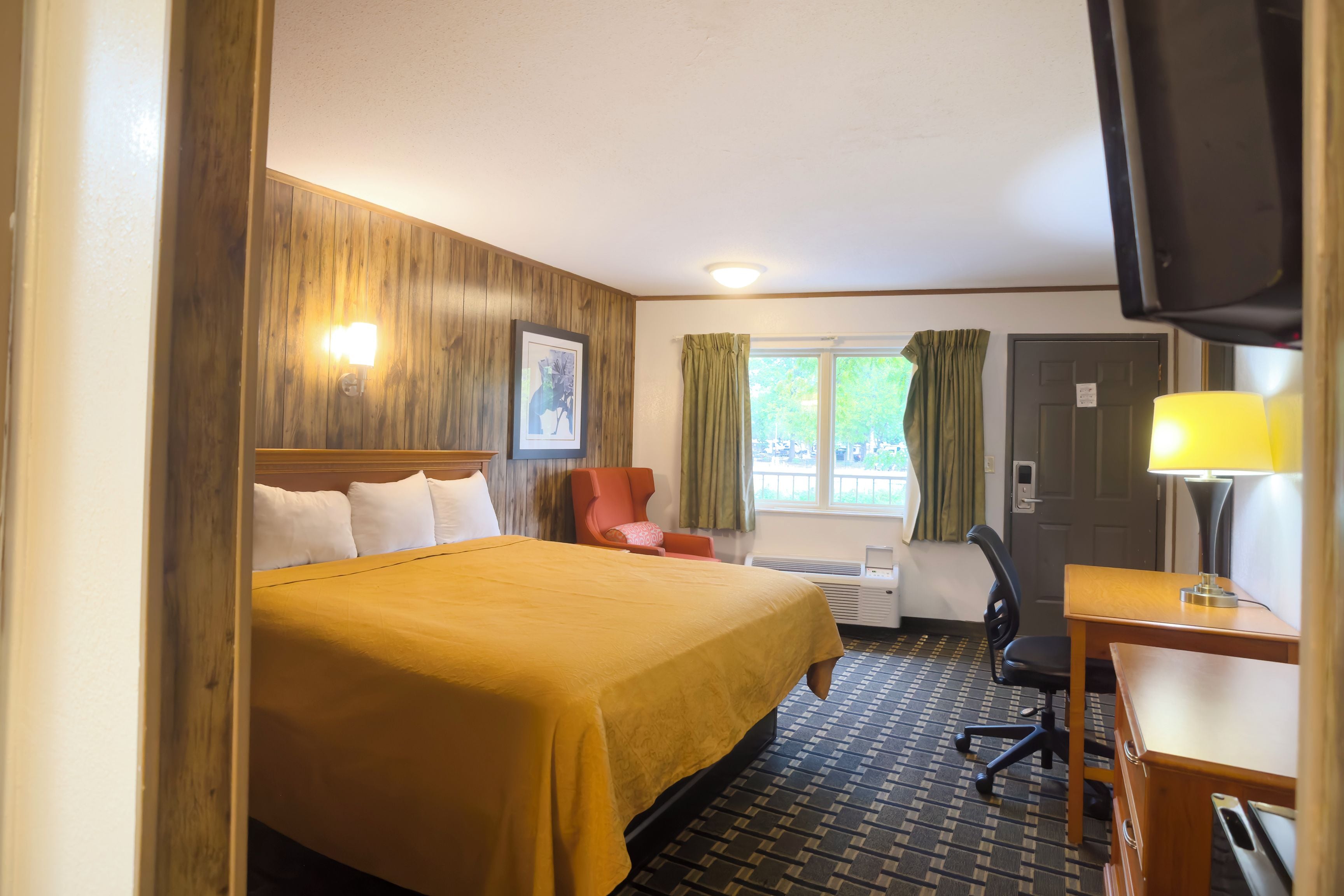 Una habitación de hotel con cama, escritorio, silla, televisor y ventana con cortinas.