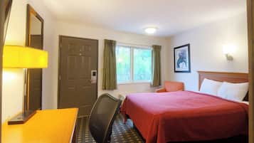 A hotel room with a bed, a desk, a chair, a door, a window with curtains, and a wall-mounted lamp.