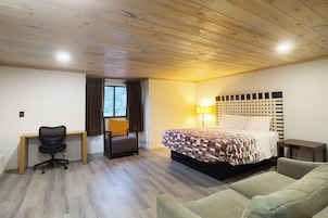 A hotel room with a wooden ceiling, a bed with a patterned bedspread, a desk with a chair, a sofa, and a window with curtains.