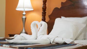 Una cama bien hecha con una toalla blanca formando un corazón, una lámpara y un cabecero de madera.
