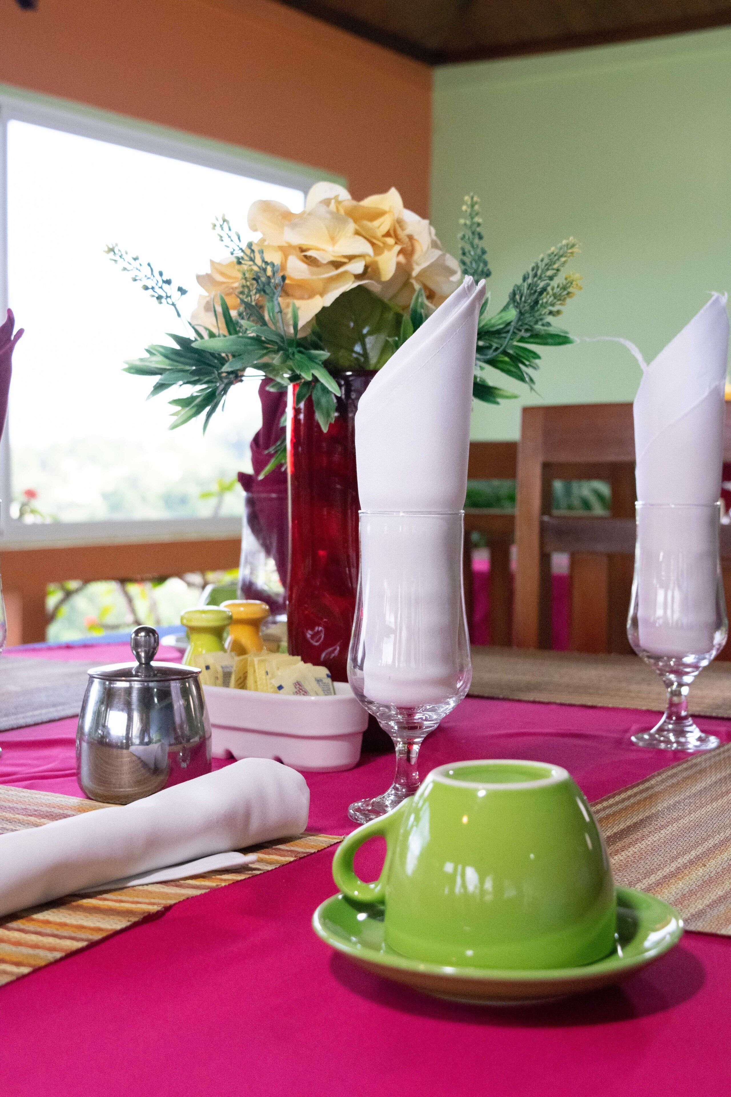 Une table dressée avec une tasse verte, des serviettes blanches, une théière en argent et un vase rempli de fleurs.