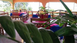 A restaurant with wooden tables and chairs, covered with blue tablecloths, and a red tablecloth at the center. - Mandeview Hotel (Mandeville)