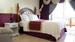 A neatly made bed with a wooden headboard, a bedside table with a lamp, and a view of the outdoors through a window with curtains. - Mandeview Hotel (Mandeville)