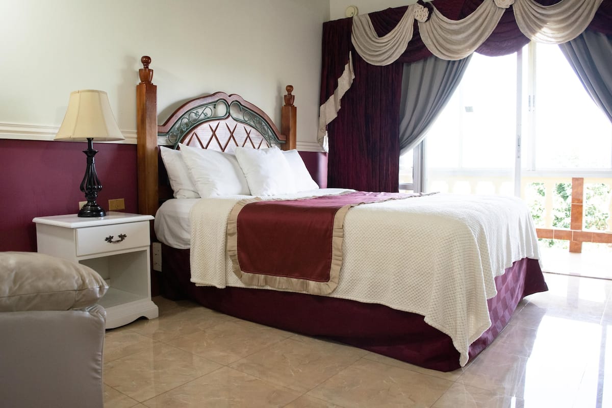 Uma cama bem arrumada com cabeceira de madeira, mesa de cabeceira com abajur, e vista do exterior por uma janela com cortinas.