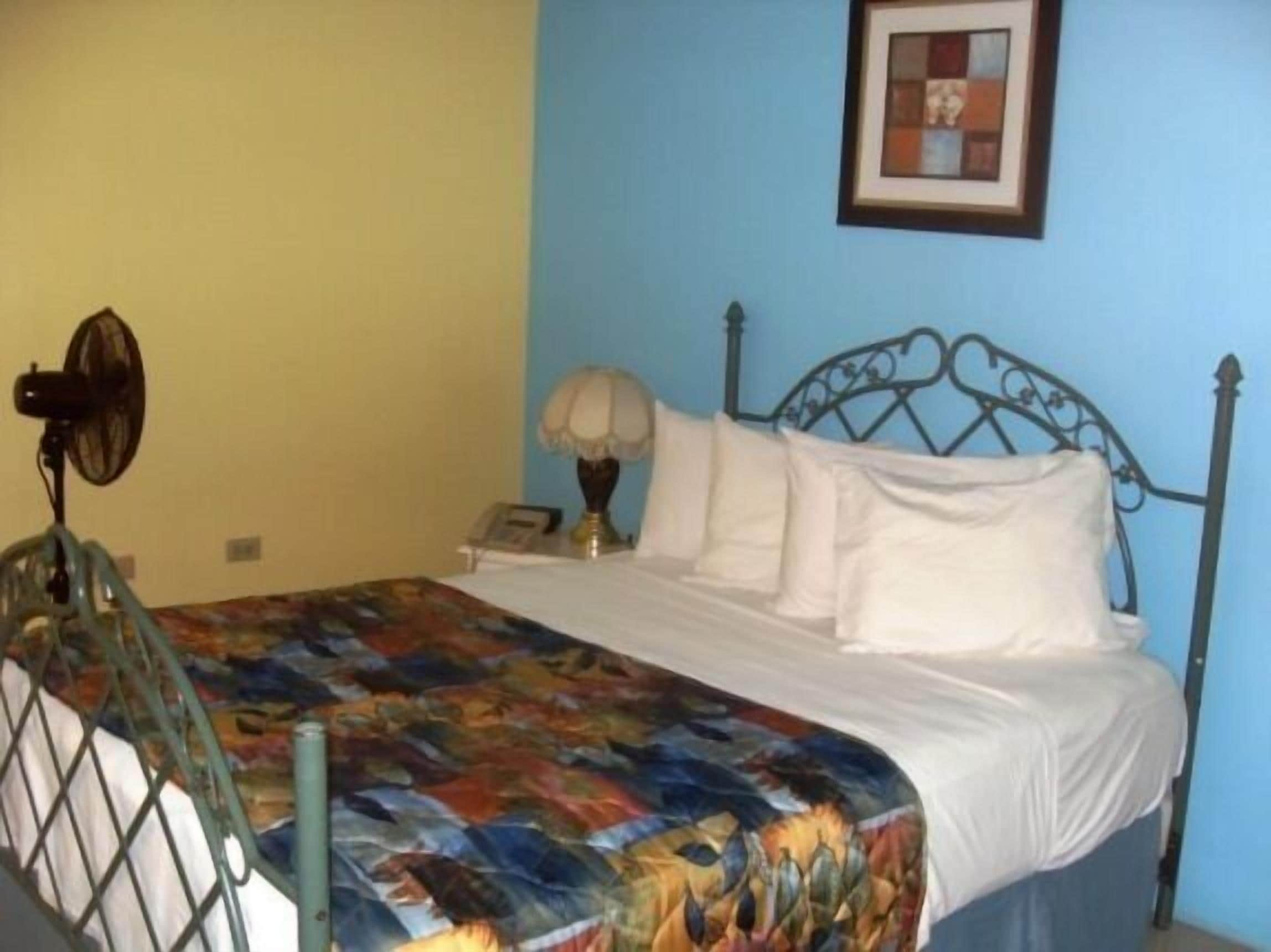 Un dormitorio con estructura de cama metálica, colcha colorida, lámpara de noche y un cuadro en la pared.