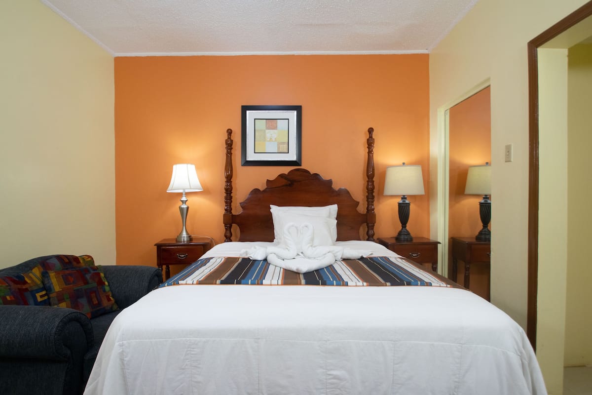 Quarto de hotel com cama, duas mesas de cabeceira com abajures, um quadro na parede e um cobertor colorido no sofá.