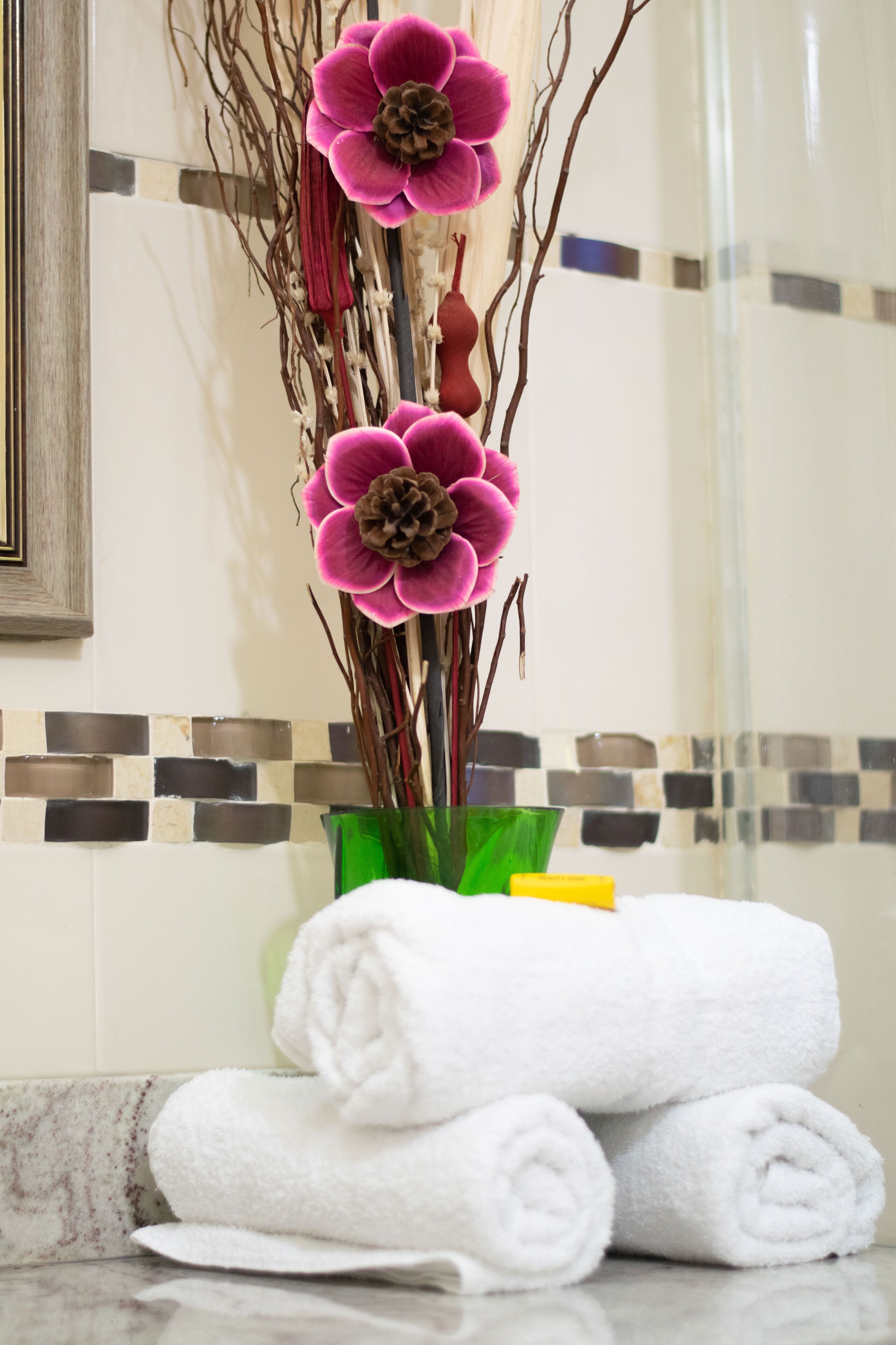Une salle de bain avec un mur recouvert de carreaux, un vase vert contenant des fleurs roses et des serviettes blanches soigneusement pliées.