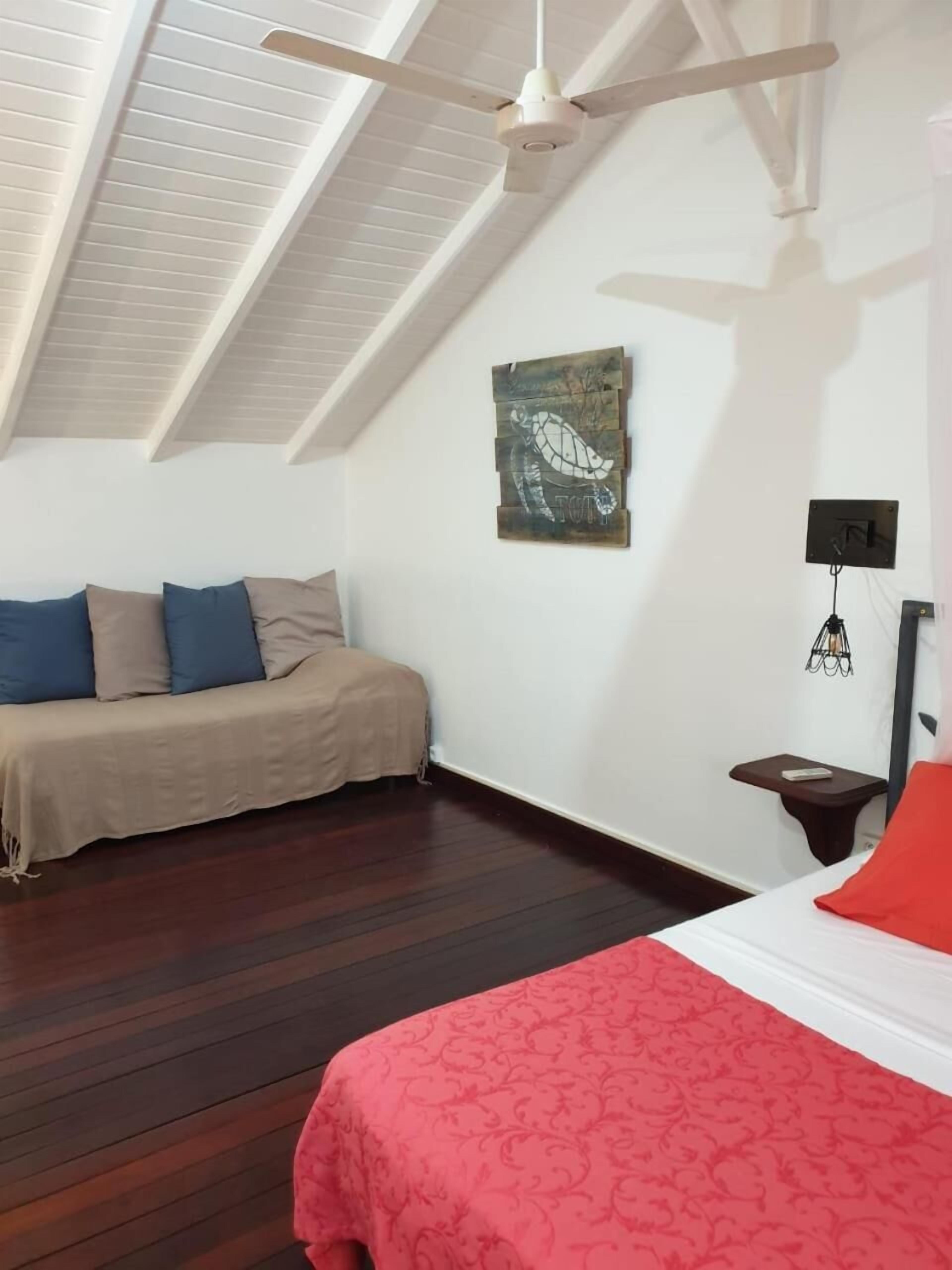 A bedroom with a sloped ceiling, a bed with a red bedspread, a sofa with beige and blue cushions, a wooden floor, and a wall-mounted artwork.