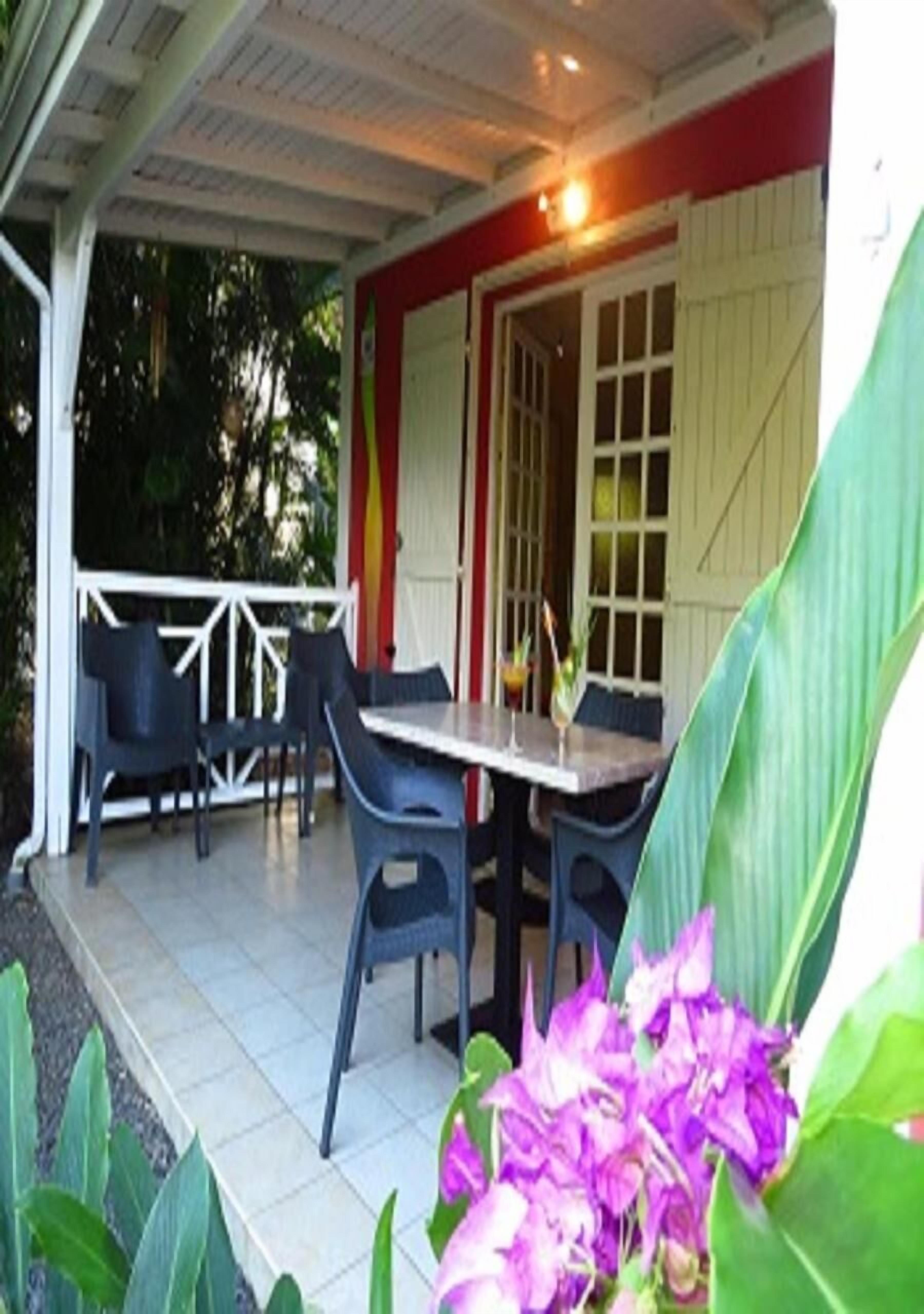 Un patio al aire libre con mesa y sillas, rodeado por una barandilla blanca y hojas verdes grandes.