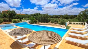 Zona de piscina amb gandules i para-sols de fibra, envoltada de vegetació i un cel serè.