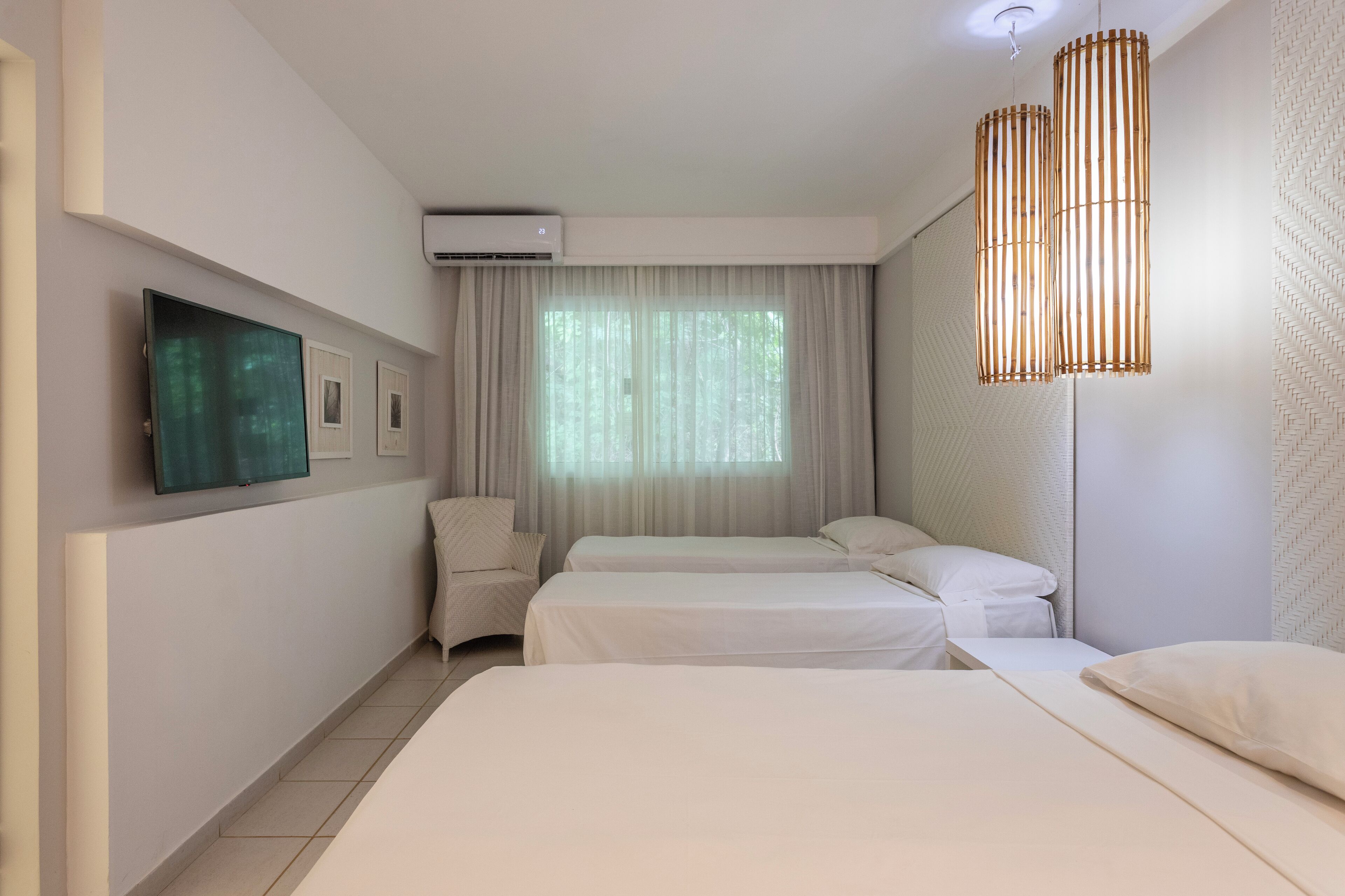 A hotel room with two beds, a flat-screen TV, an air conditioning unit, and a large window with sheer curtains.