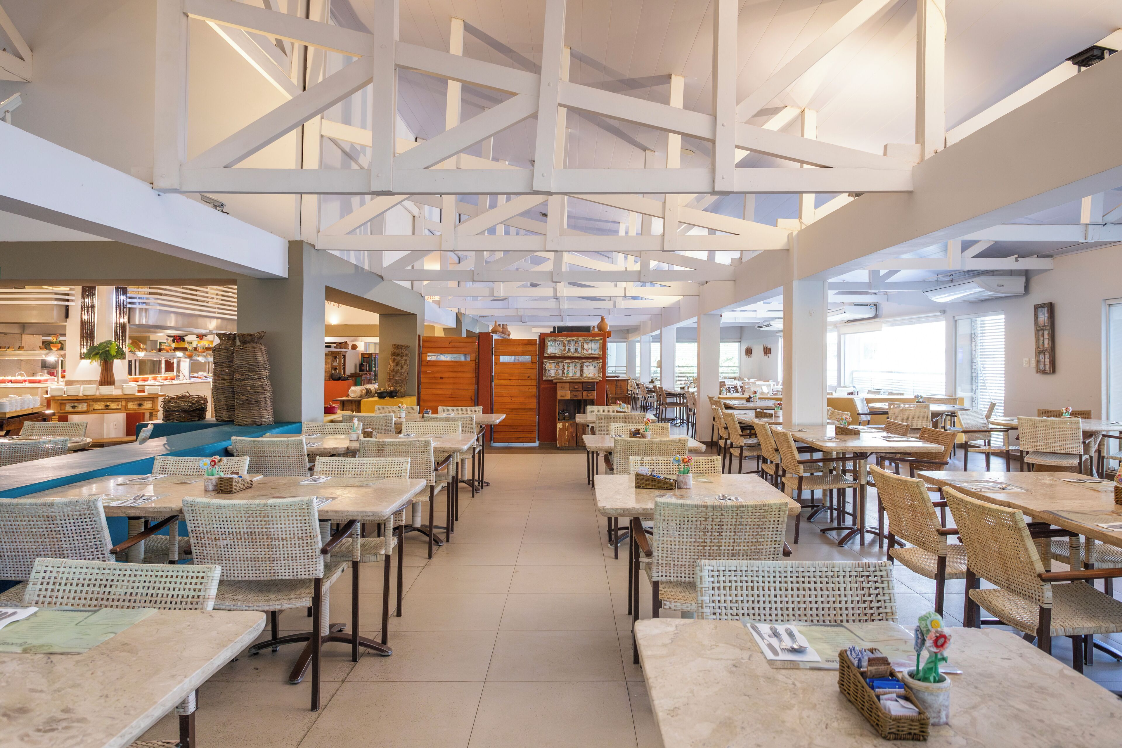 A spacious restaurant with high ceilings and white beams, featuring a variety of seating arrangements and a central kitchen area.