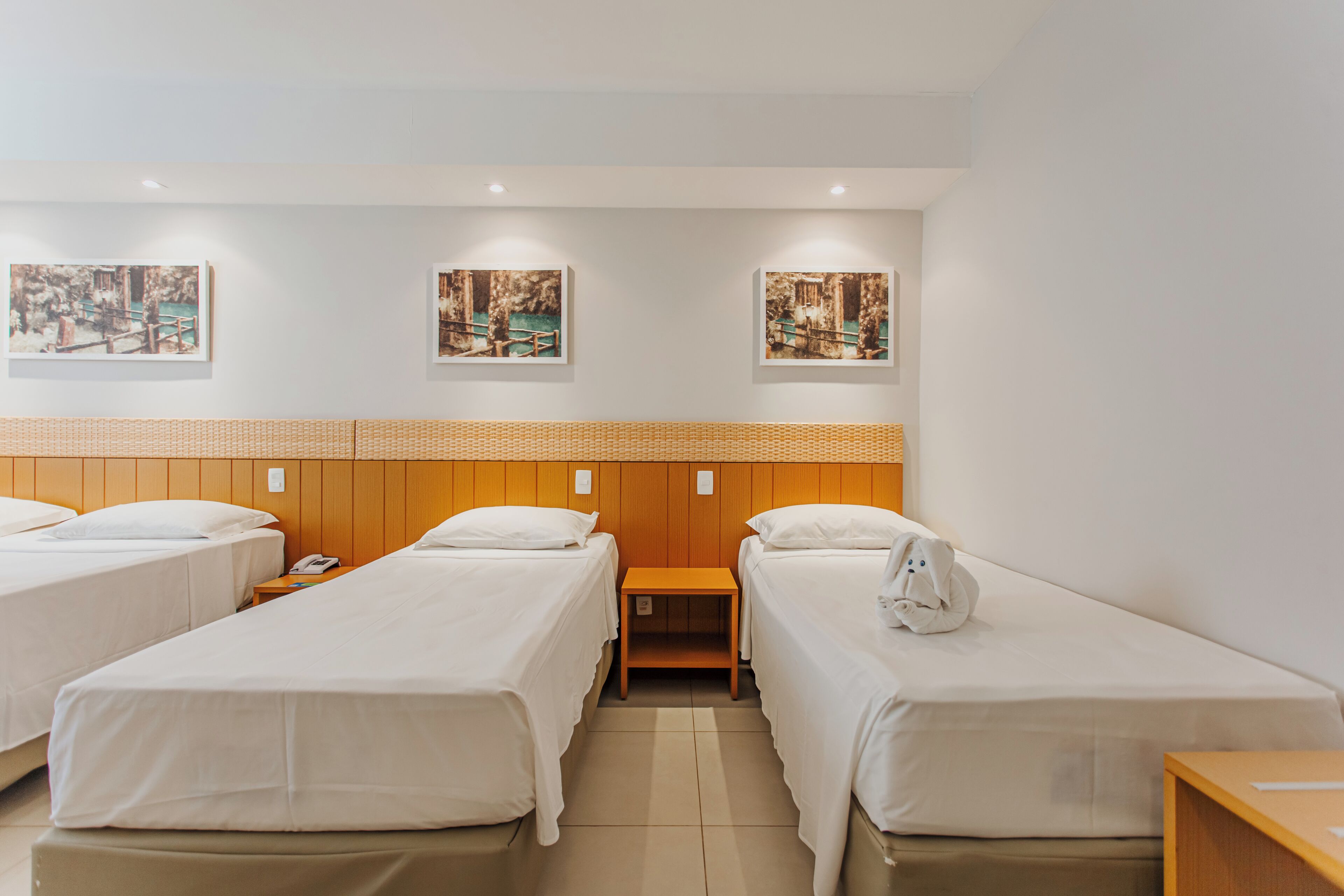 A hotel room with two beds, a wooden headboard, a small table, and three framed pictures on the wall.