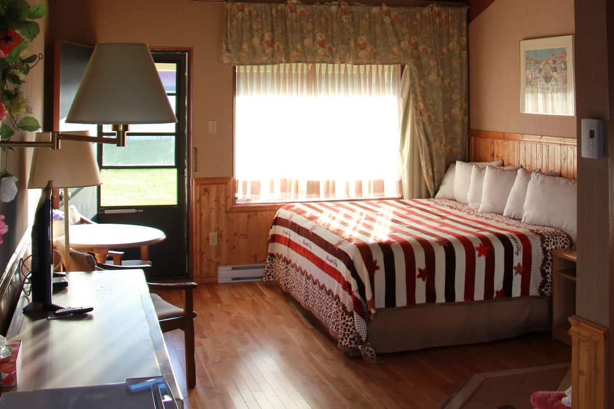 A room with a wooden ceiling, a bed with a striped blanket, a desk with a lamp, and a window with curtains.