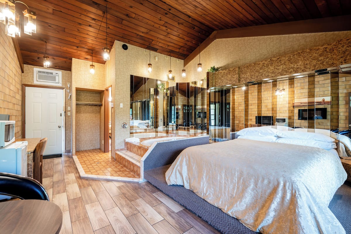 A bedroom with a large bed, wooden ceiling, and a bathroom with a shower and a mirror.