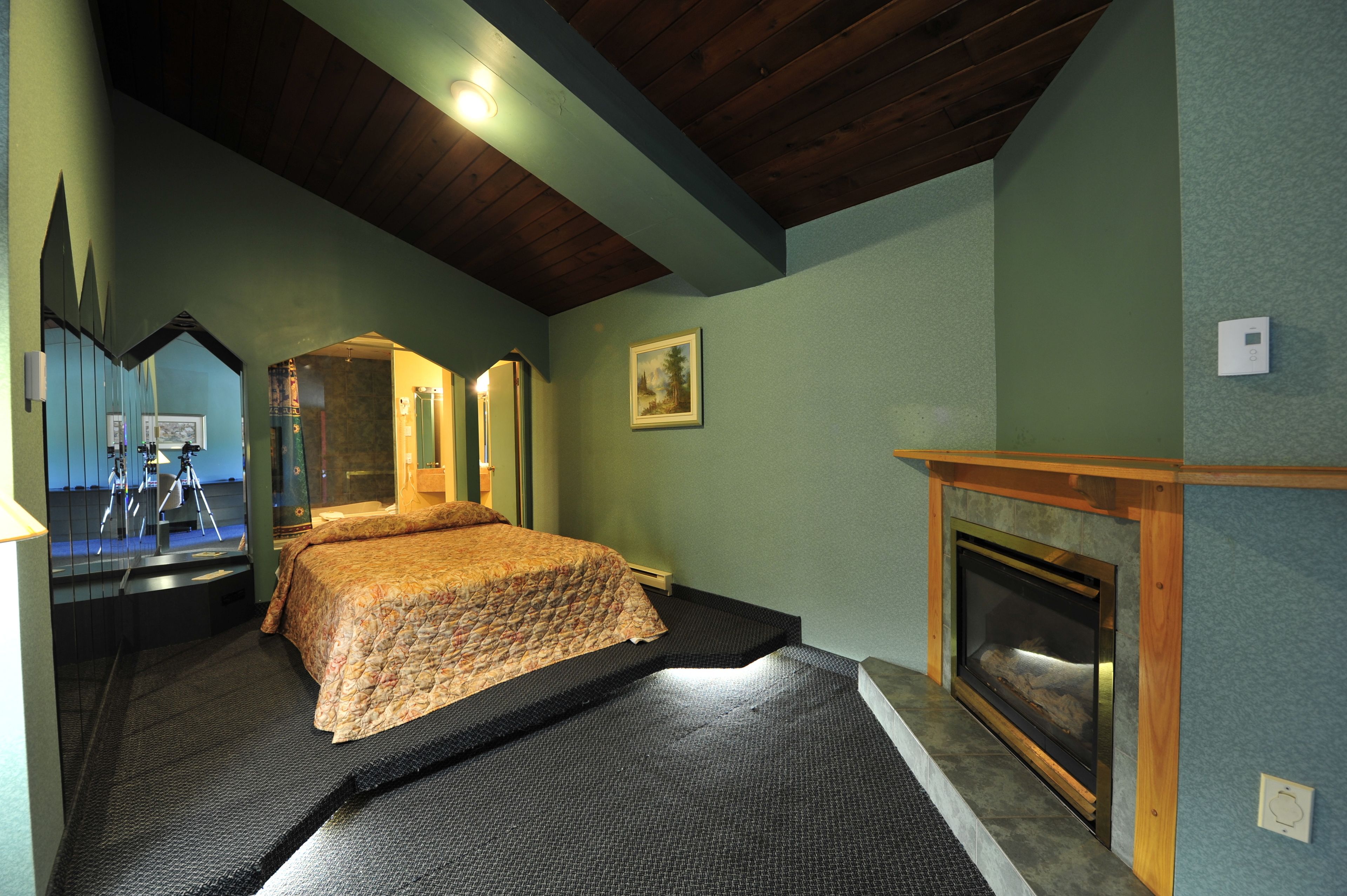A bedroom with a bed, a fireplace, a mirror, and a ceiling with wooden beams.