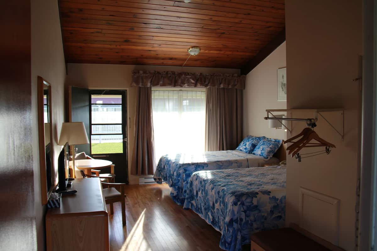 A hotel room with two beds, a desk with a lamp, a chair, a window with curtains, and a coat rack.