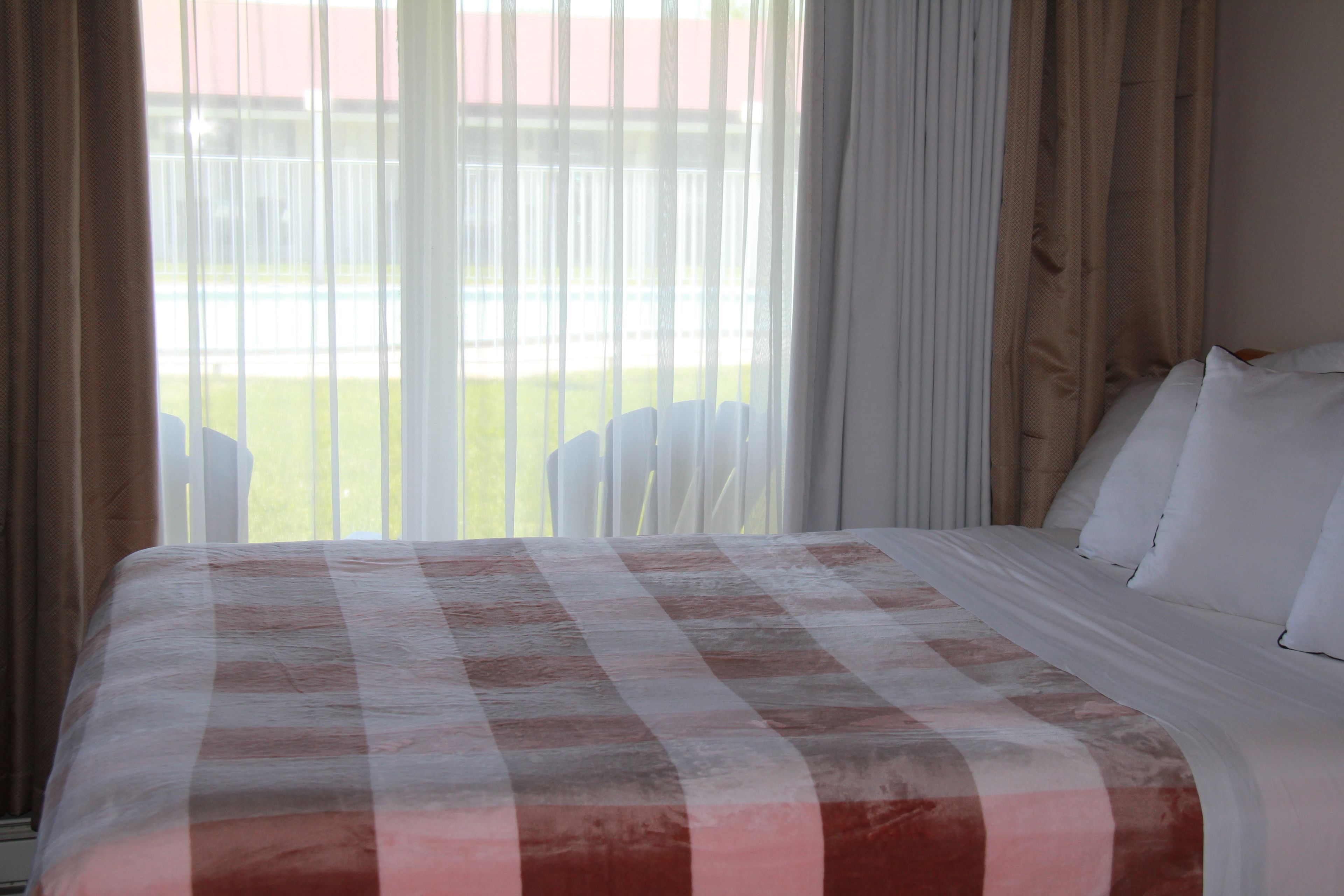 A hotel room with a large bed, white curtains, and a view of a pool and lawn.