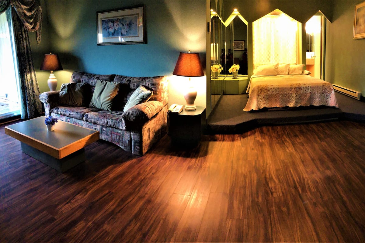 A hotel room with a sofa, a small table, a bedside table with a lamp, a bed with a canopy, and a mirror.