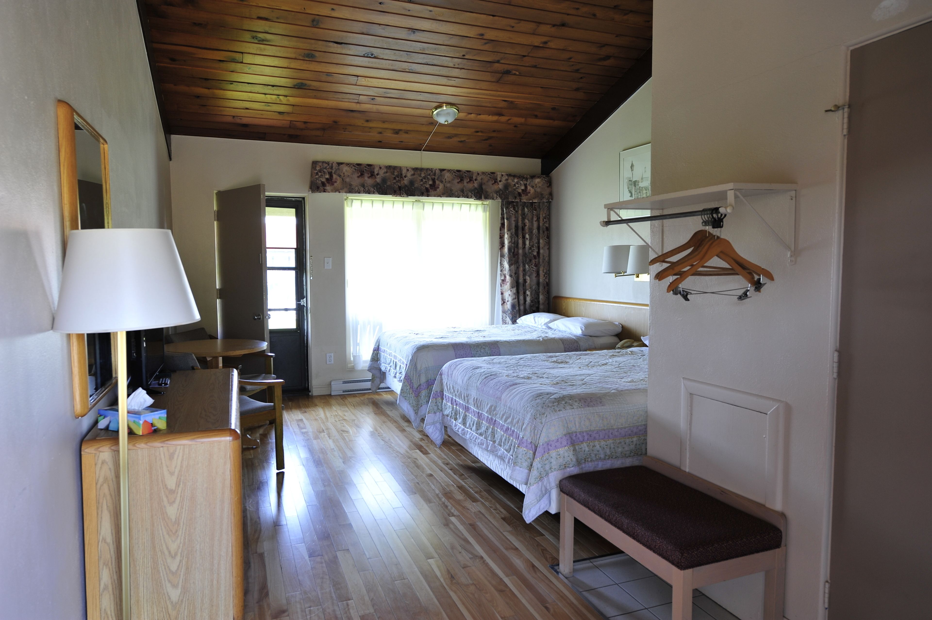 Una habitación con dos camas, un escritorio con lámpara, un banco, techo de madera y una puerta que da a otra habitación.