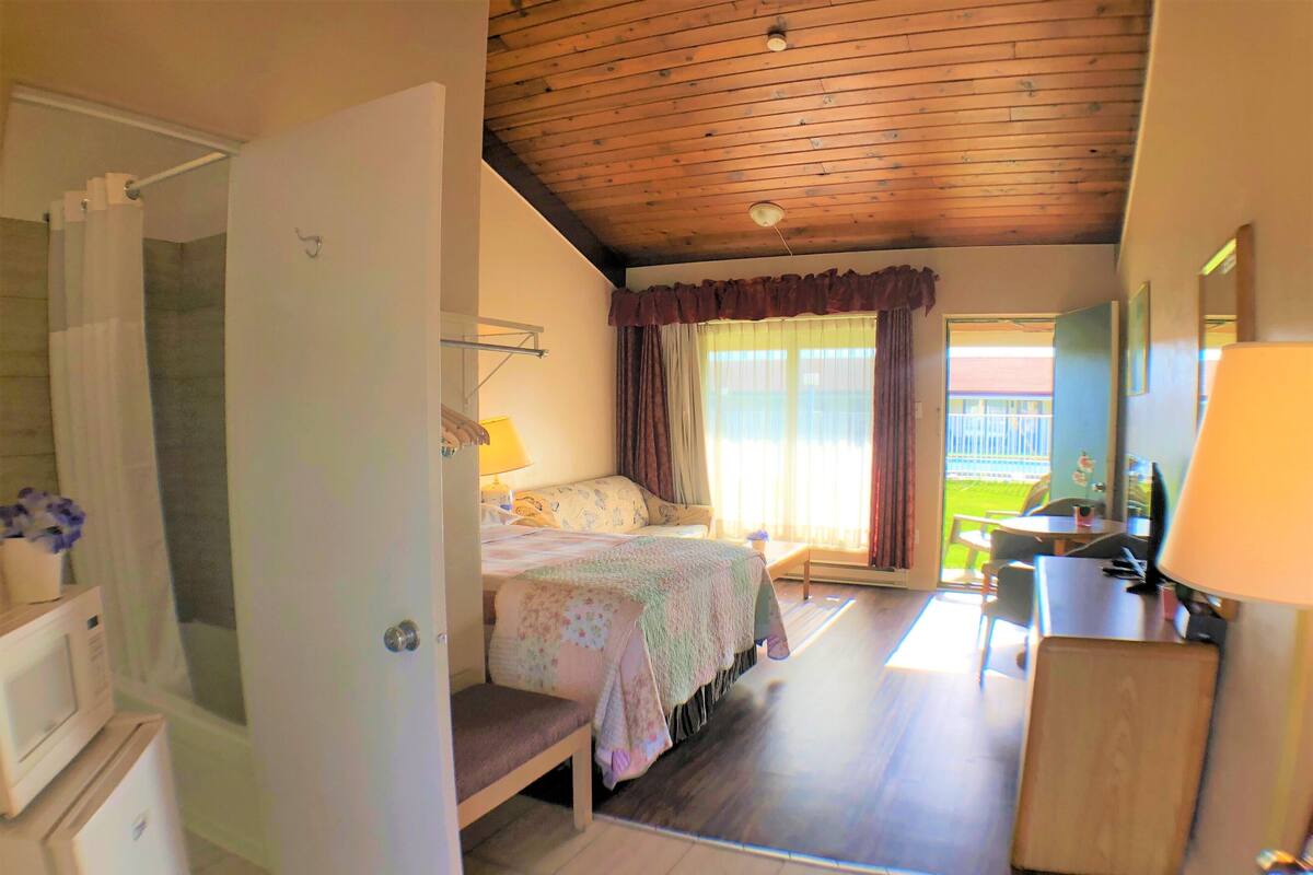 A bedroom with a bed, a desk, a chair, a lamp, and a view of the outdoors.