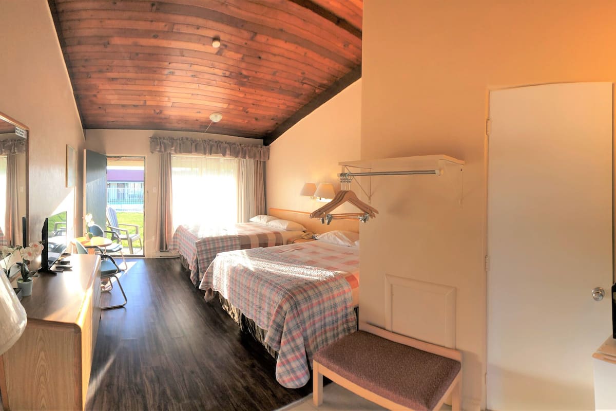 A hotel room with a wooden ceiling, two beds with plaid bedding, a desk with a lamp, a microwave, and a view of the outdoors.