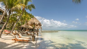 A beach with lounge chairs, palm trees, and a thatched-roof hut. - Beachfront Hotel La Palapa - Adults Only (Isla Holbox)
