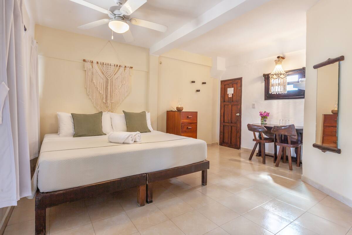 Quarto com cama, mesa de cabeceira de madeira, escrivaninha com cadeiras, ventilador de teto e lustre.