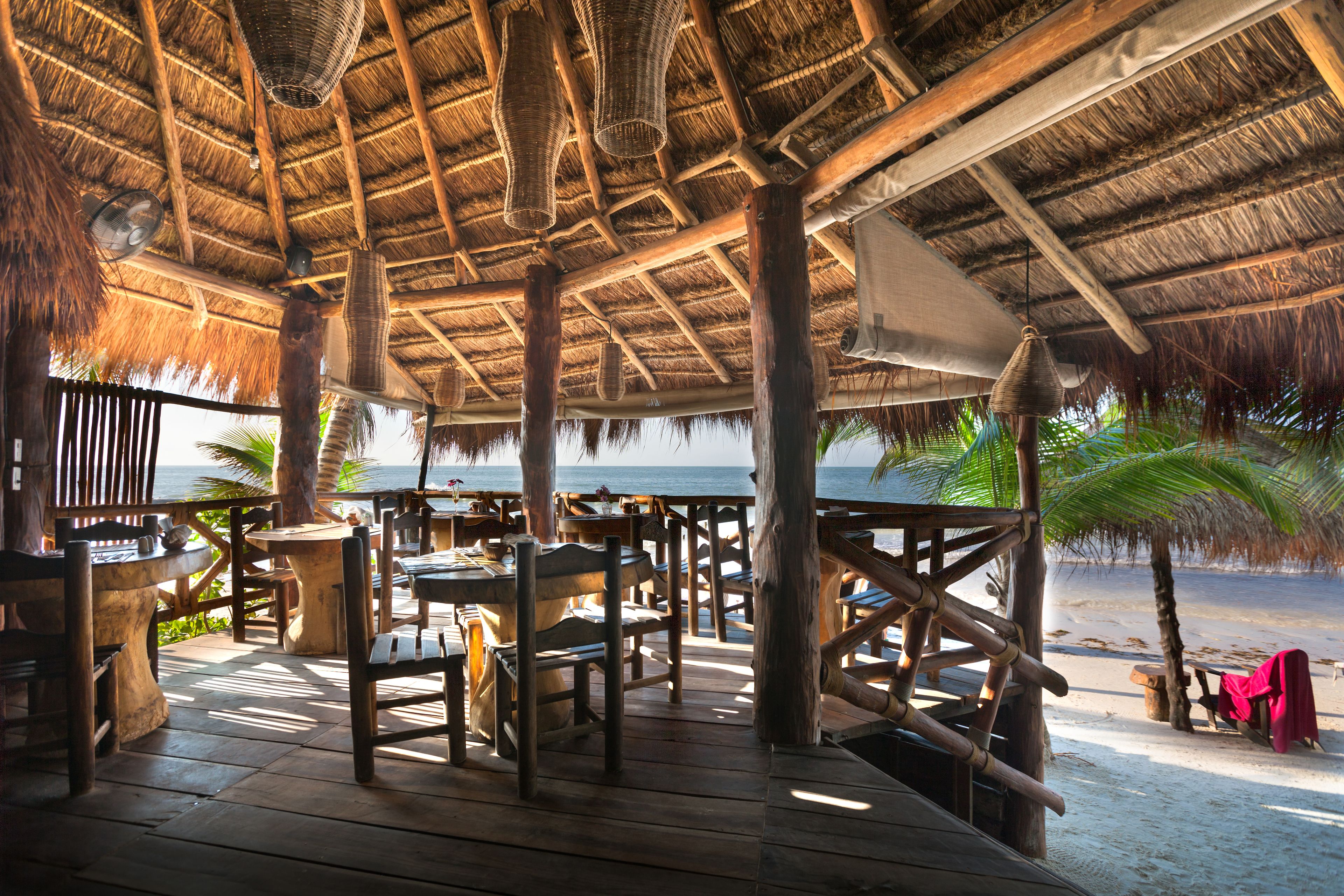 Une terrasse extérieure en bois, avec des tables et des chaises, un toit de chaume et une vue sur la plage.