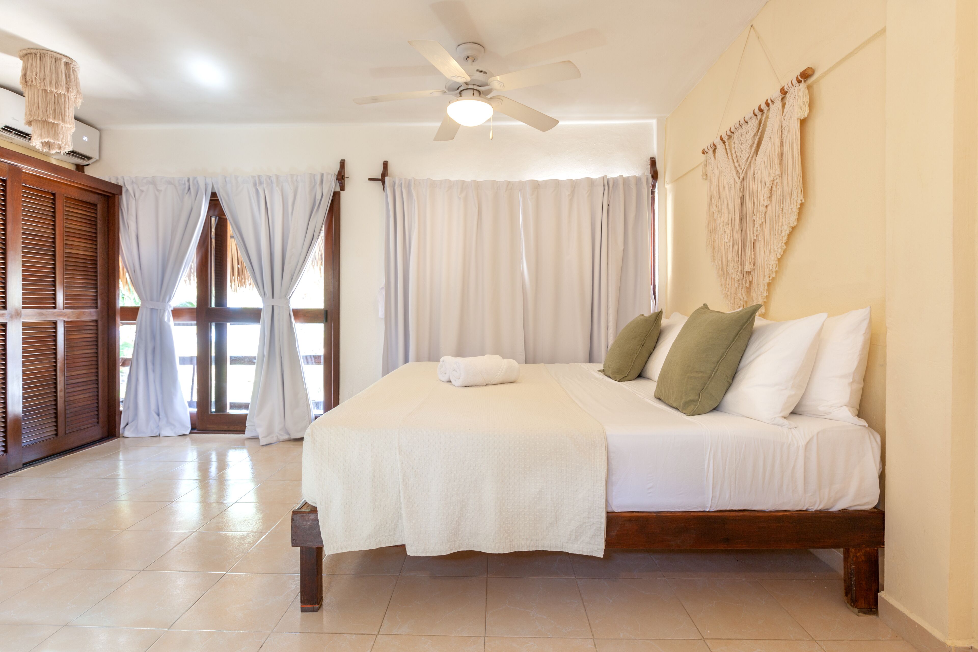 A bedroom with a bed, curtains, a ceiling fan, and a wall-mounted macrame piece.