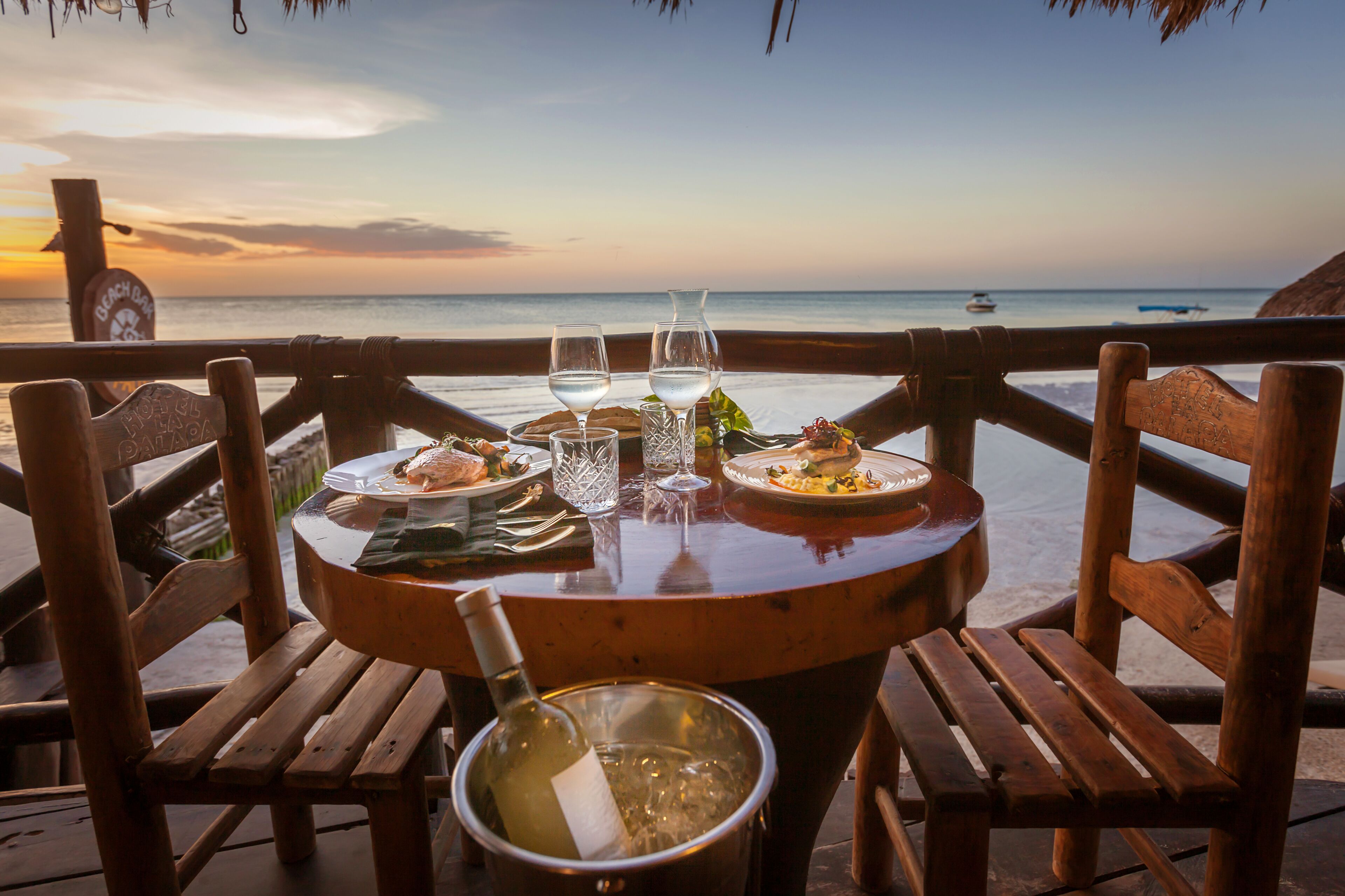 A wooden table with a bottle of wine, two glasses, and plates of food set against a view of the ocean at sunset.
