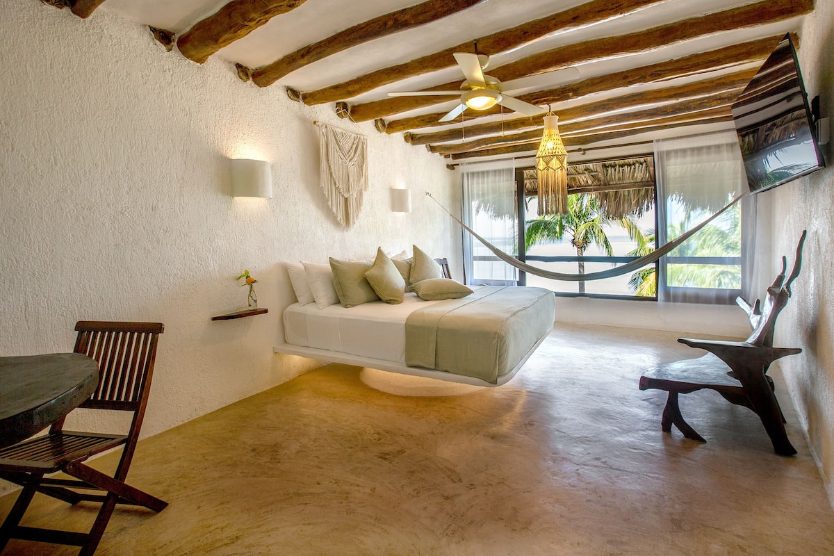 Quarto com teto de madeira, um sofá branco com almofadas, uma mesa de jantar de madeira e uma rede.