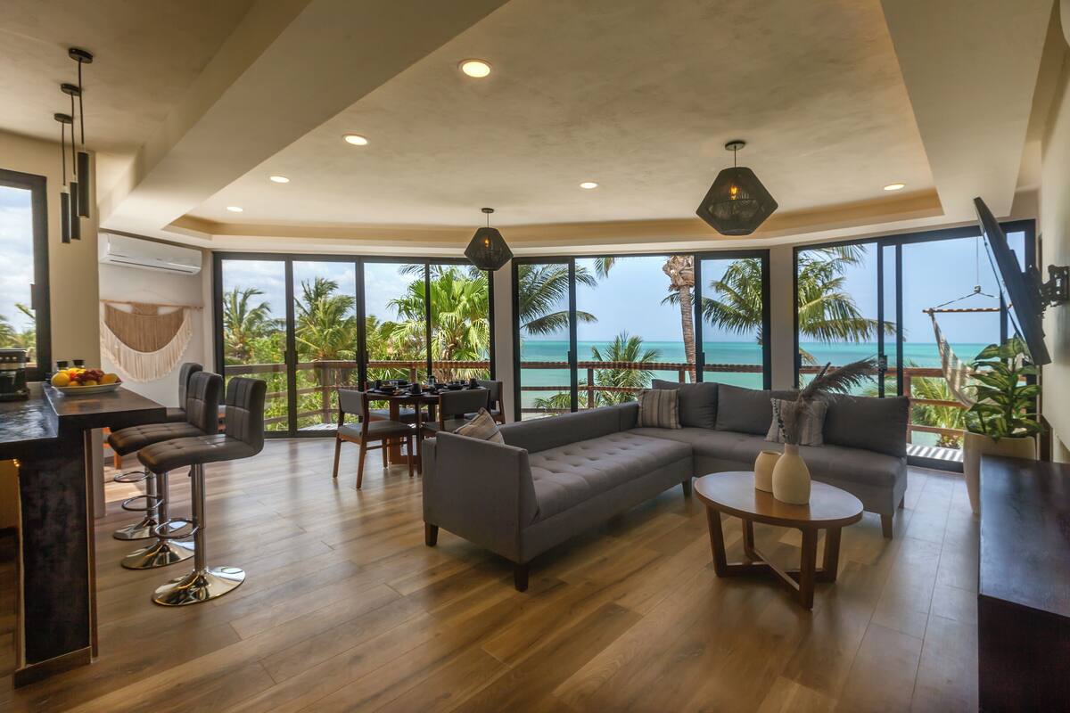 Uma sala de estar ampla com um sofá grande, uma mesa de jantar e banquetas de bar. Há vista para o oceano através de janelas grandes.