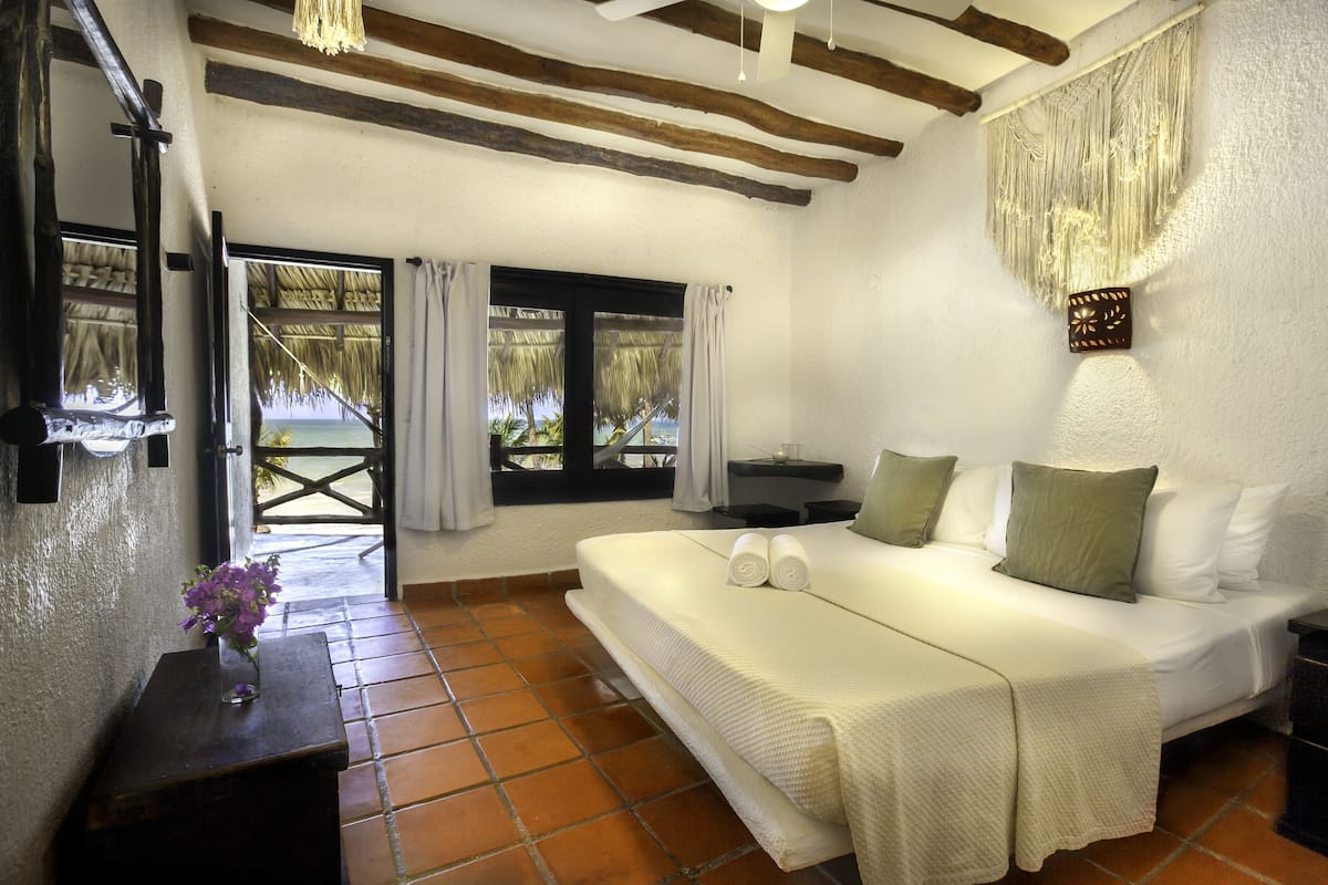 Quarto com cama, almofadas, mesa de cabeceira, um vaso com flores e vista para o exterior.