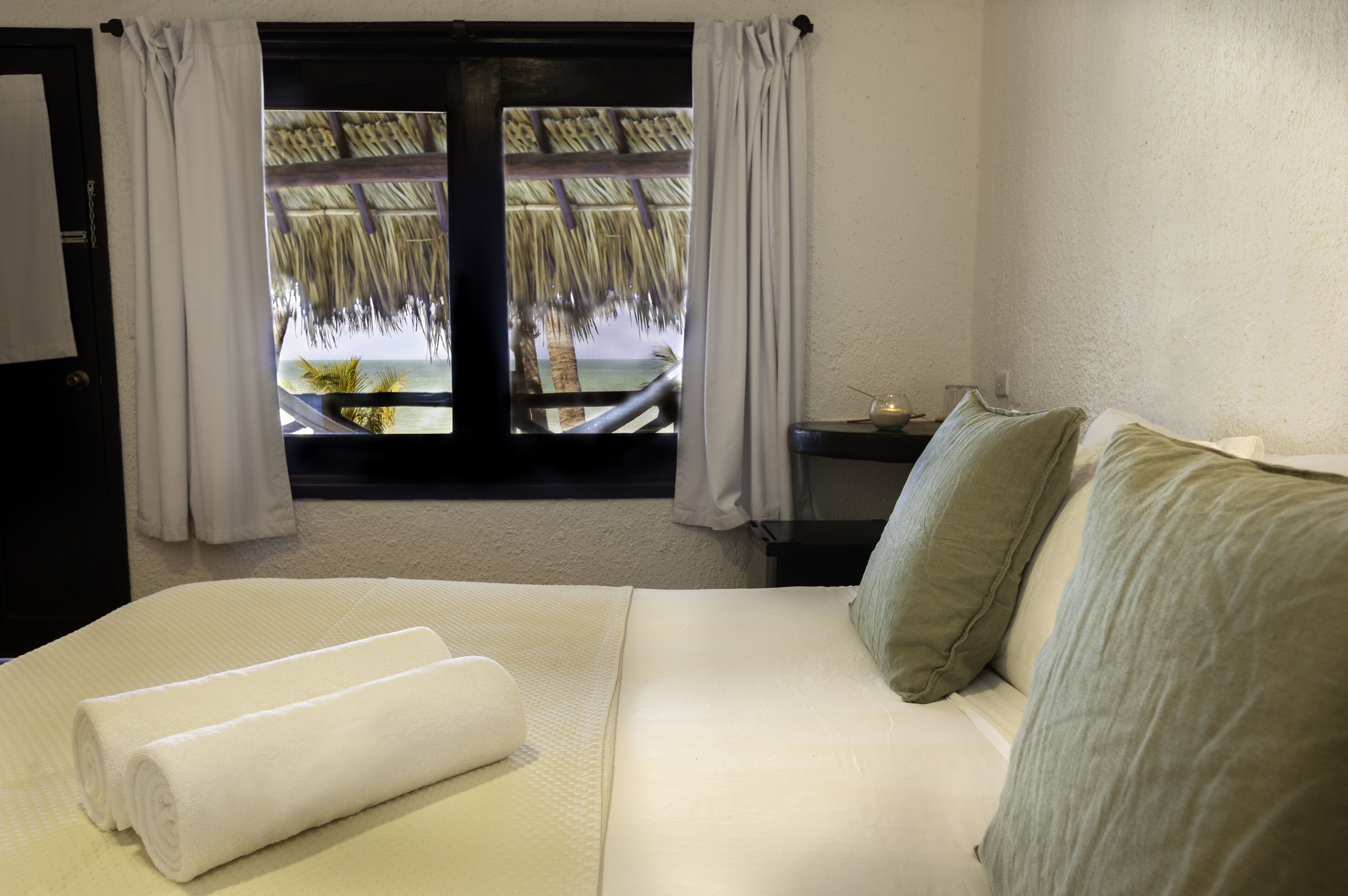 Une chambre d’hôtel avec un lit, une table de chevet et une fenêtre donnant sur un toit de chaume et des palmiers.