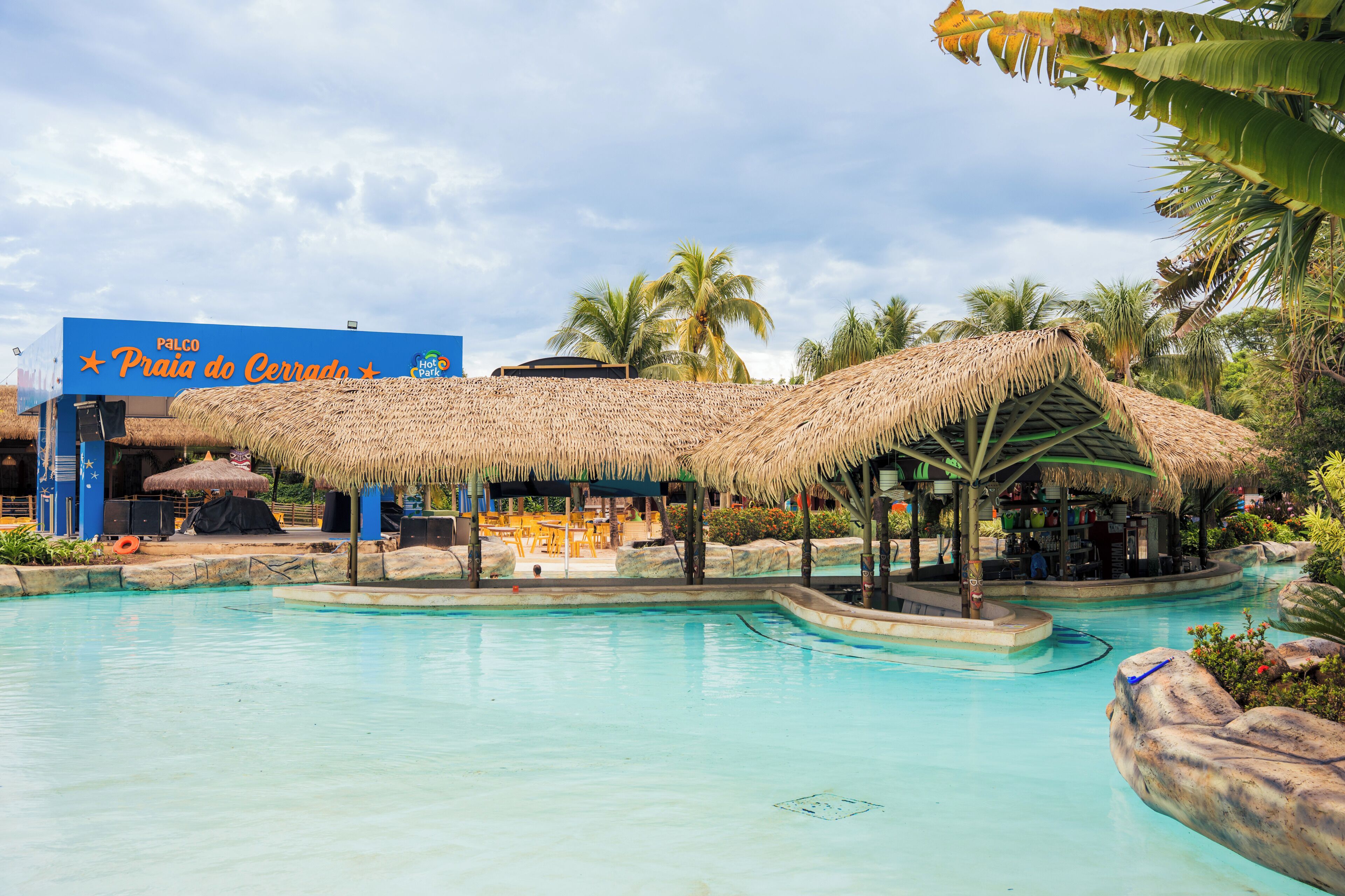 A pool area with a thatched-roof structure, palm trees, and a sign for 'Praia do Cerrado.'
