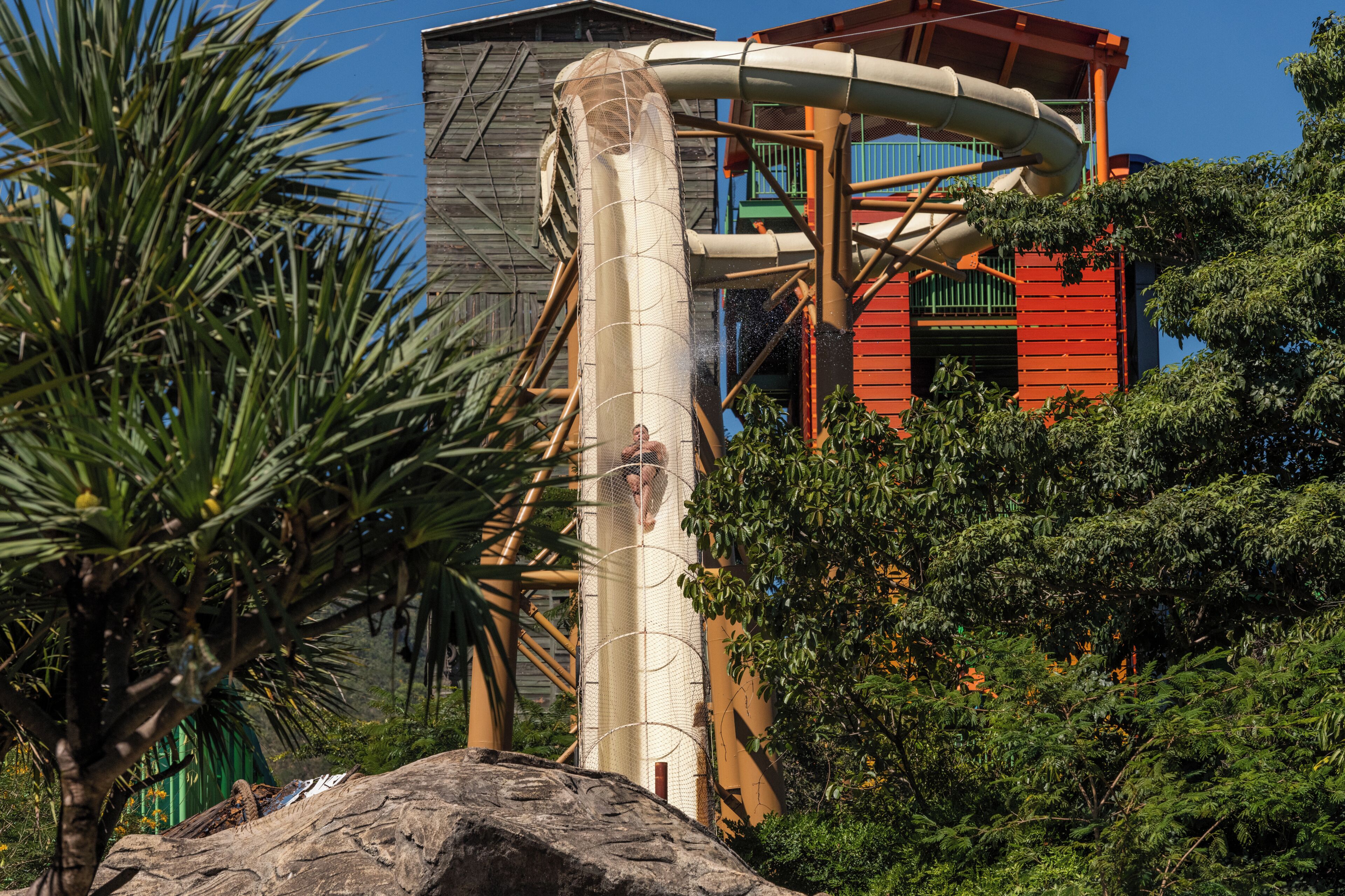 A water slide structure with a netted enclosure, a red building, and surrounding greenery.