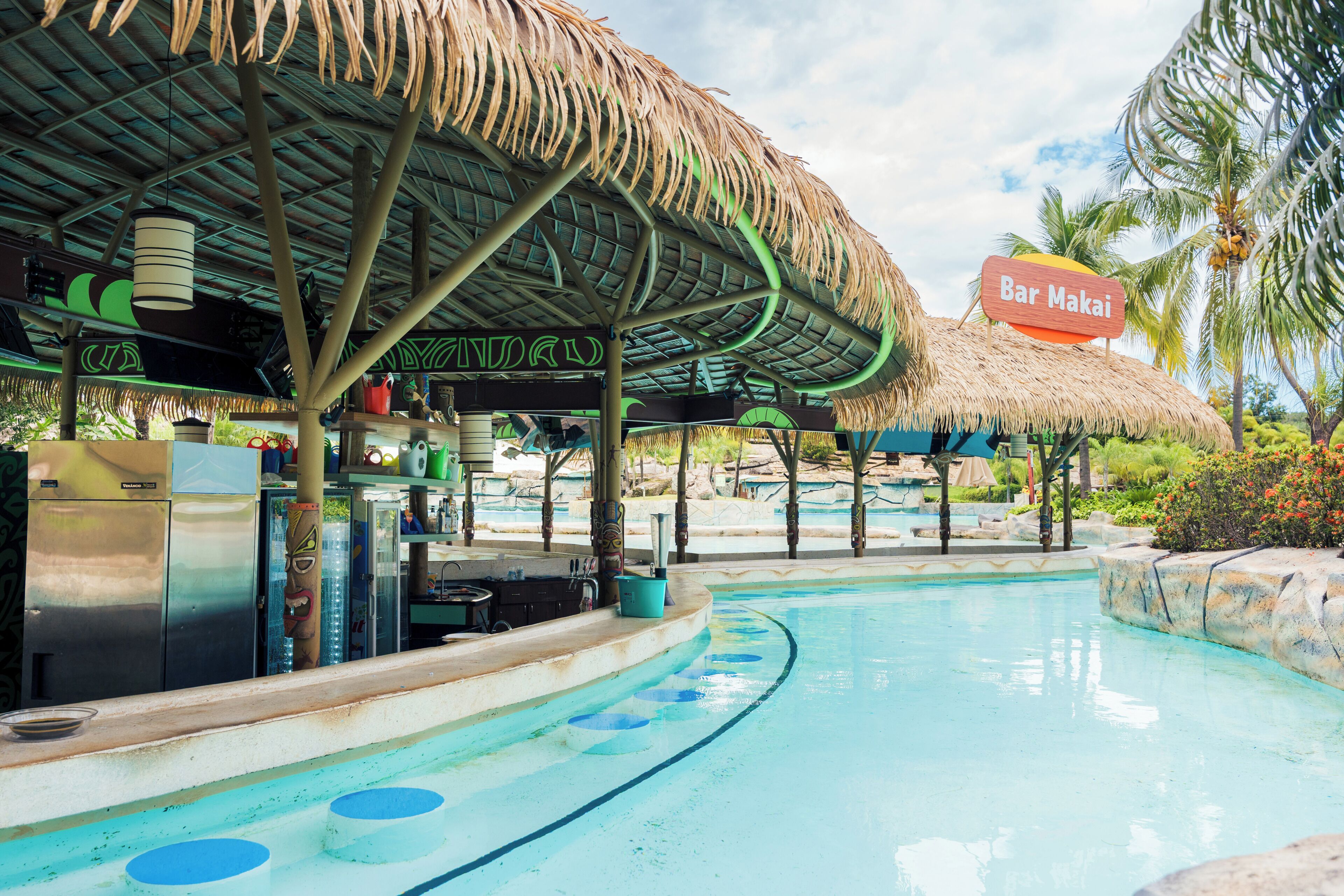 Uma área de piscina com um bar chamado 'Bar Makai', uma estrutura com telhado de palha e um cenário tropical.