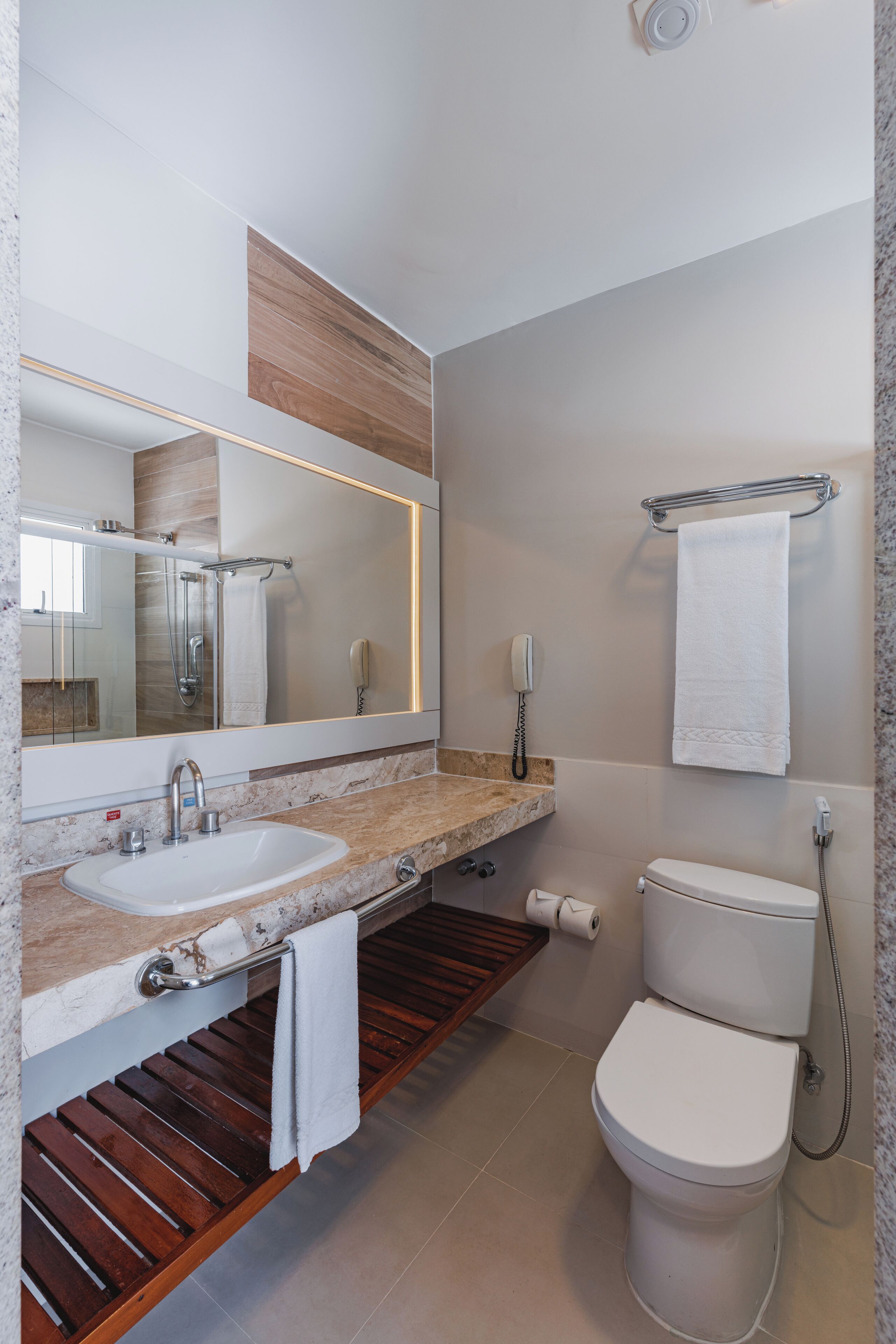 Un baño moderno con lavamanos de mármol, tocador de madera, un espejo grande, inodoro y perchero para toallas.