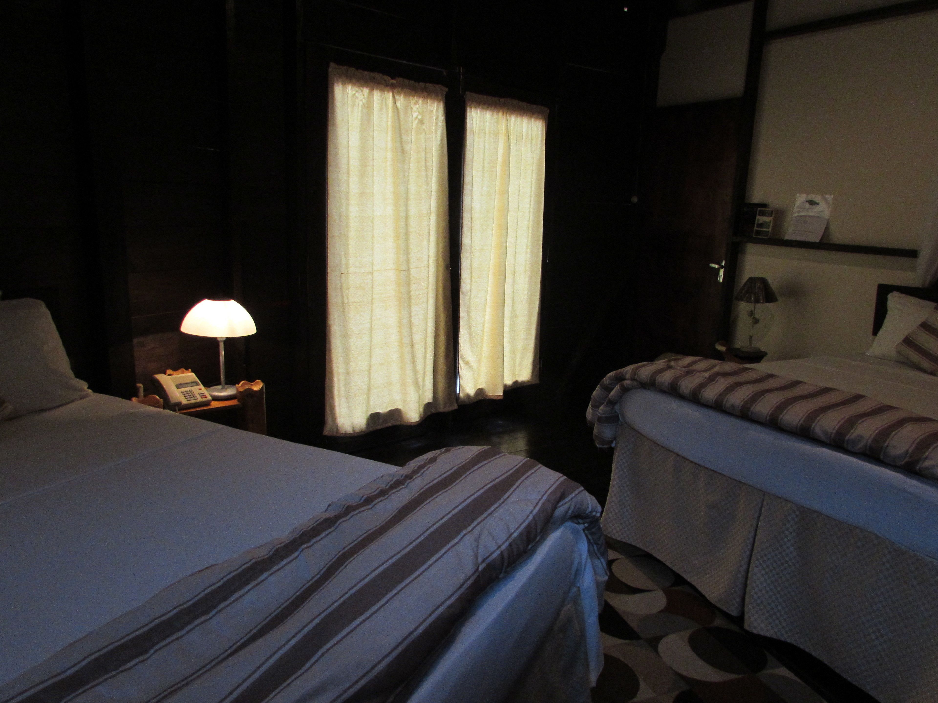 Una camera d'albergo con due letti, un comodino con una lampada e una finestra con tende.